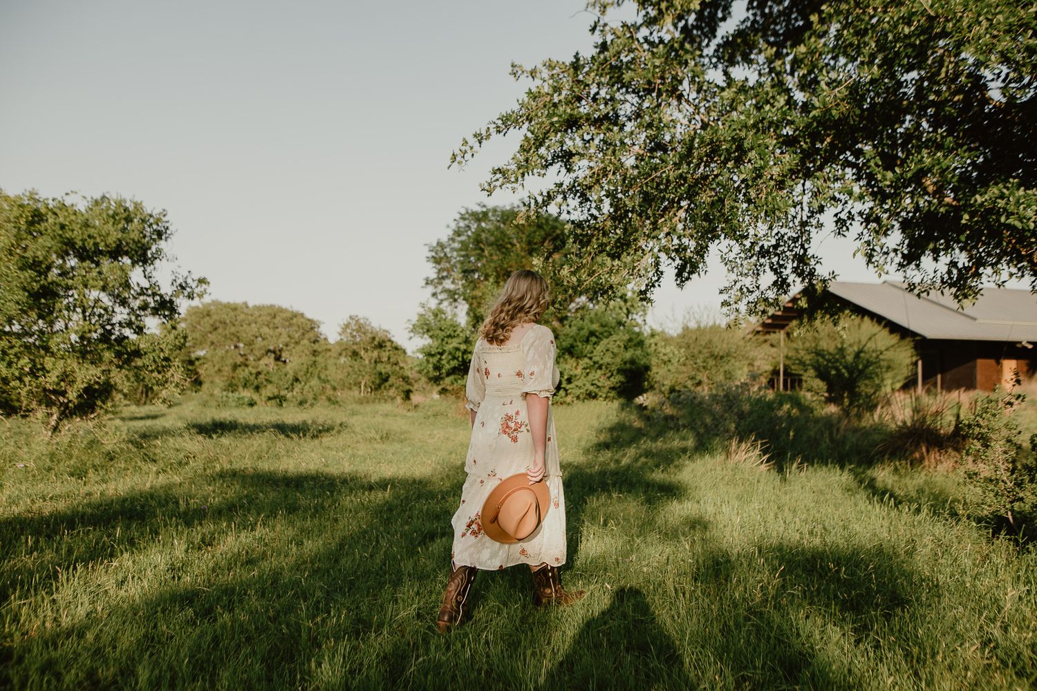 Person in floral dress sits in old wooden wheelbarrow in sunlit green field with house in background.
