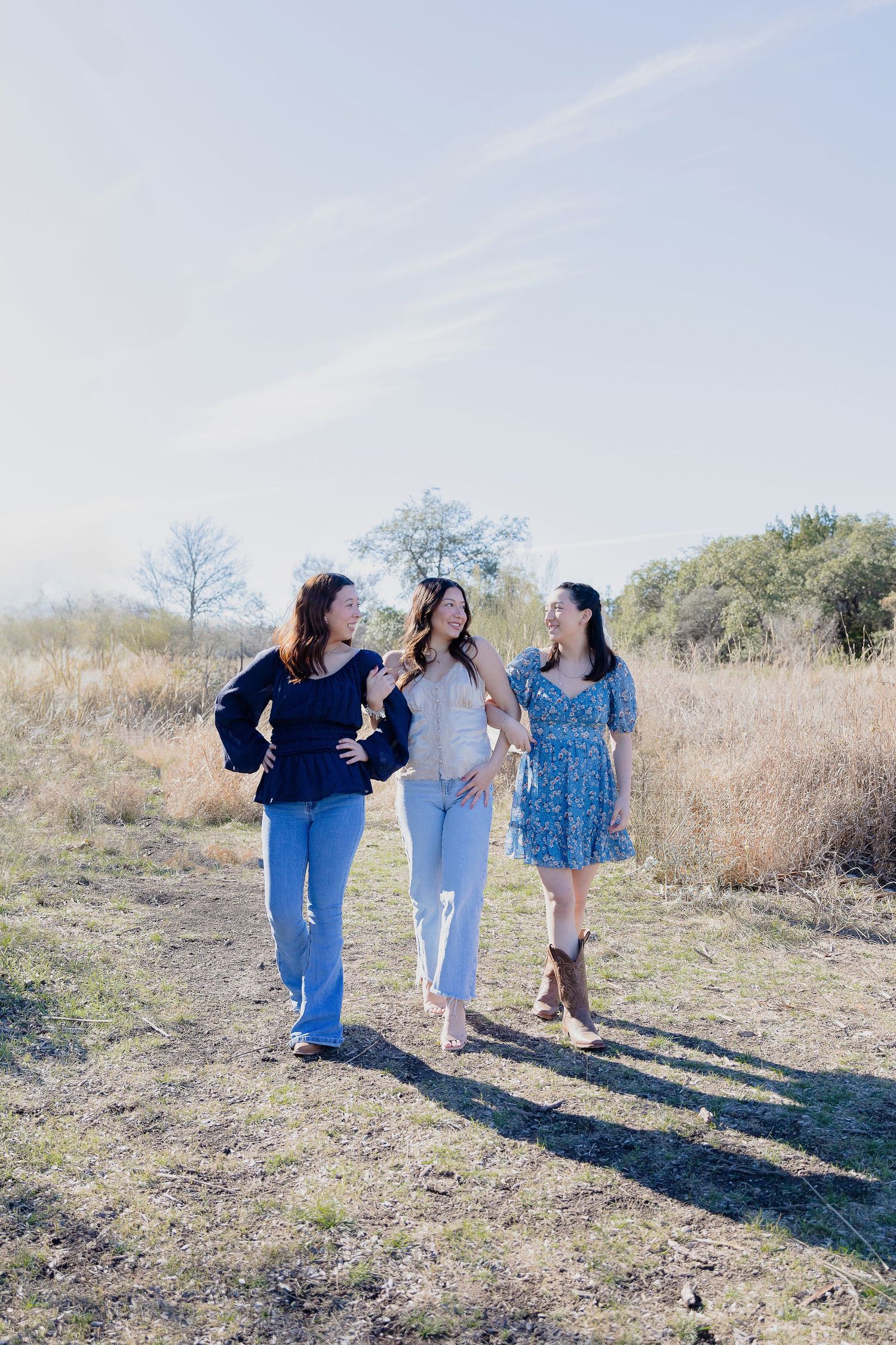 Three women walking and laughing together on a dirt path through a sunlit field on a bright autumn day.