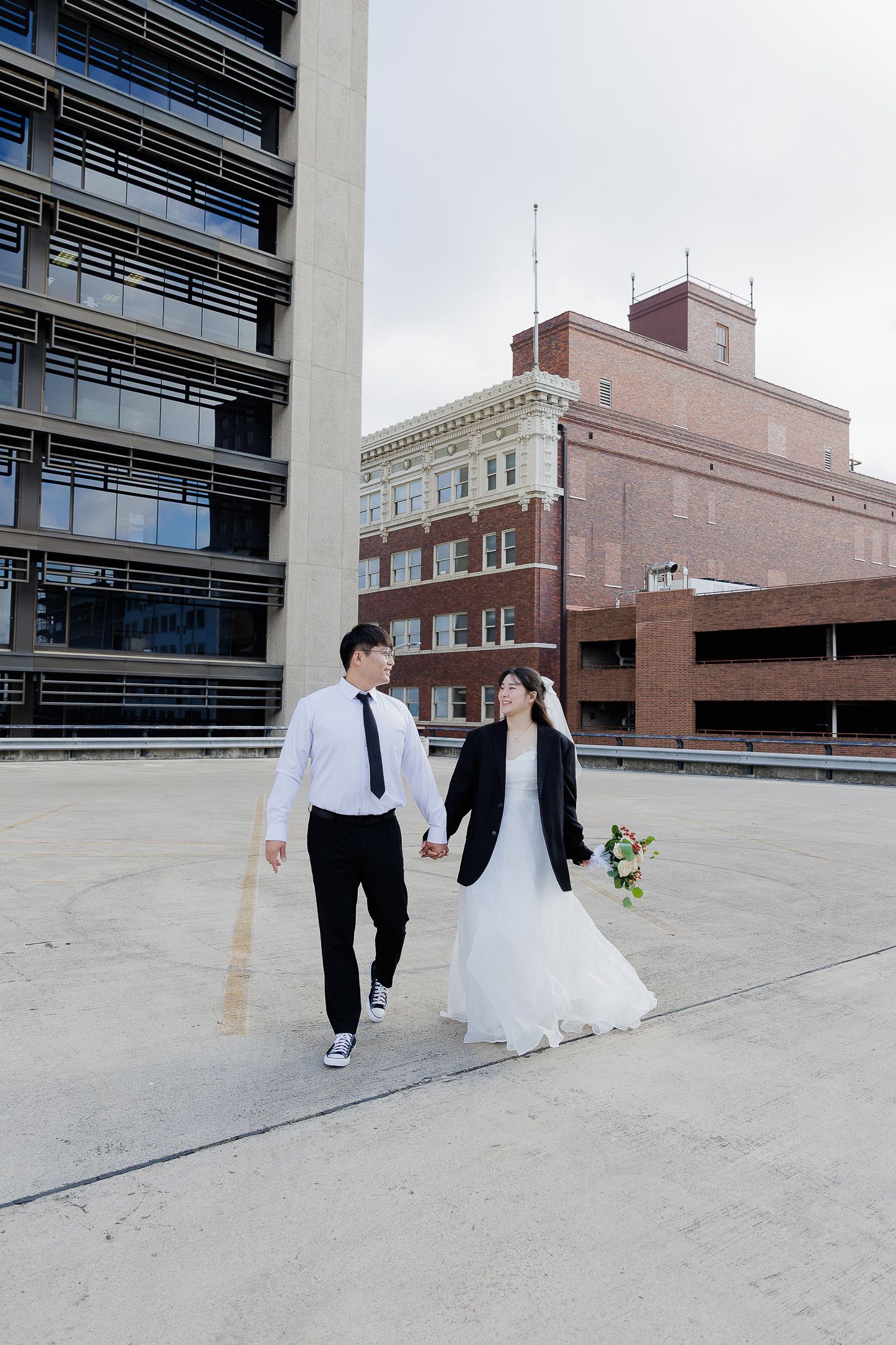A wedding couple walks across an urban rooftop parking deck with city buildings in the background.
