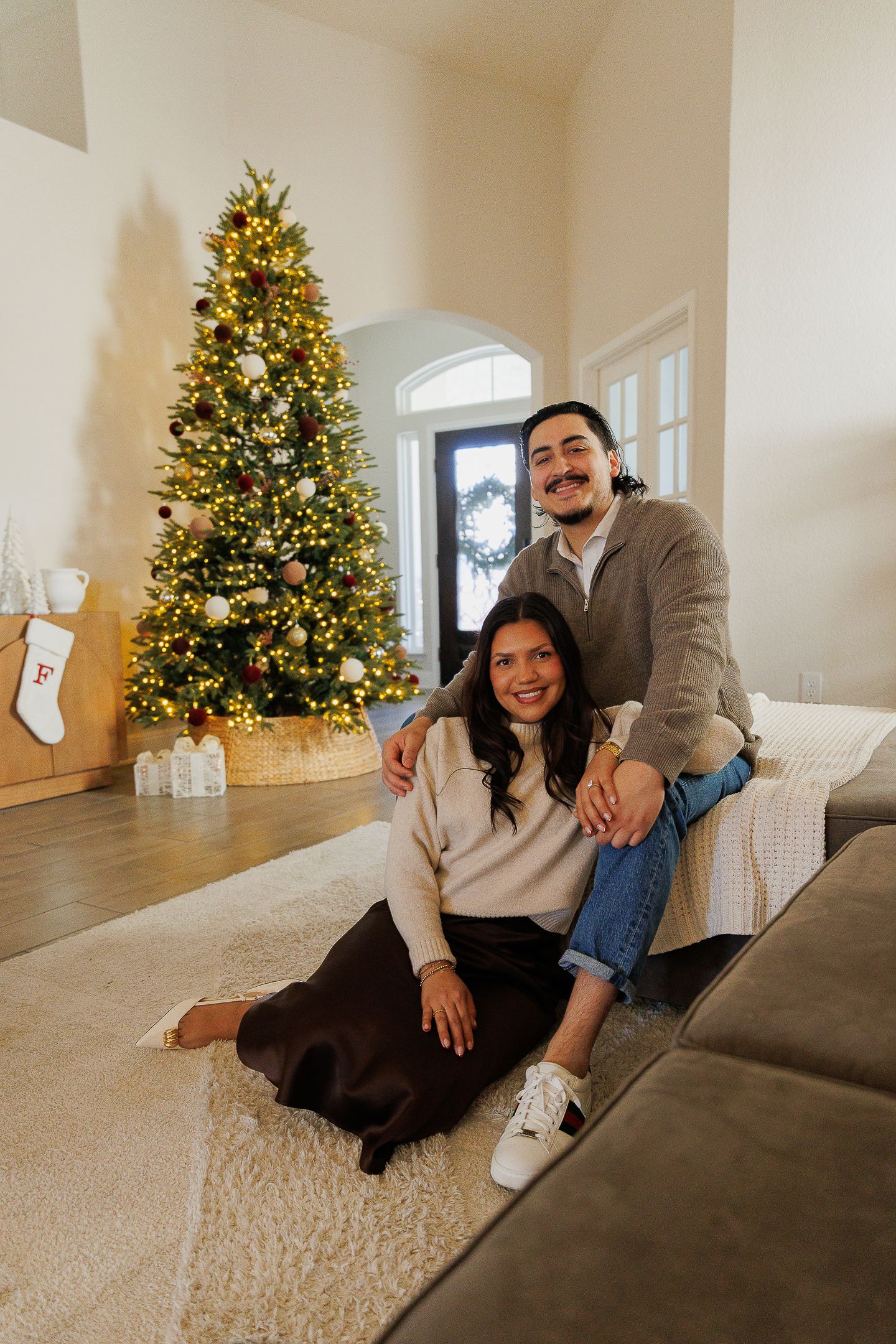 A couple sits together on a beige carpet with a decorated Christmas tree glowing in the background.