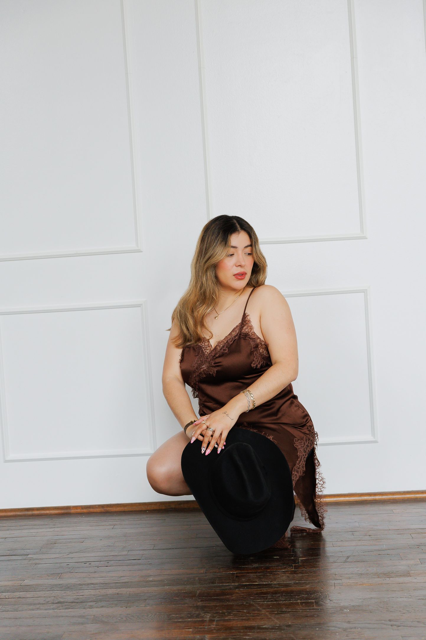 Woman in brown satin slip dress kneeling on hardwood floor against white paneled wall.