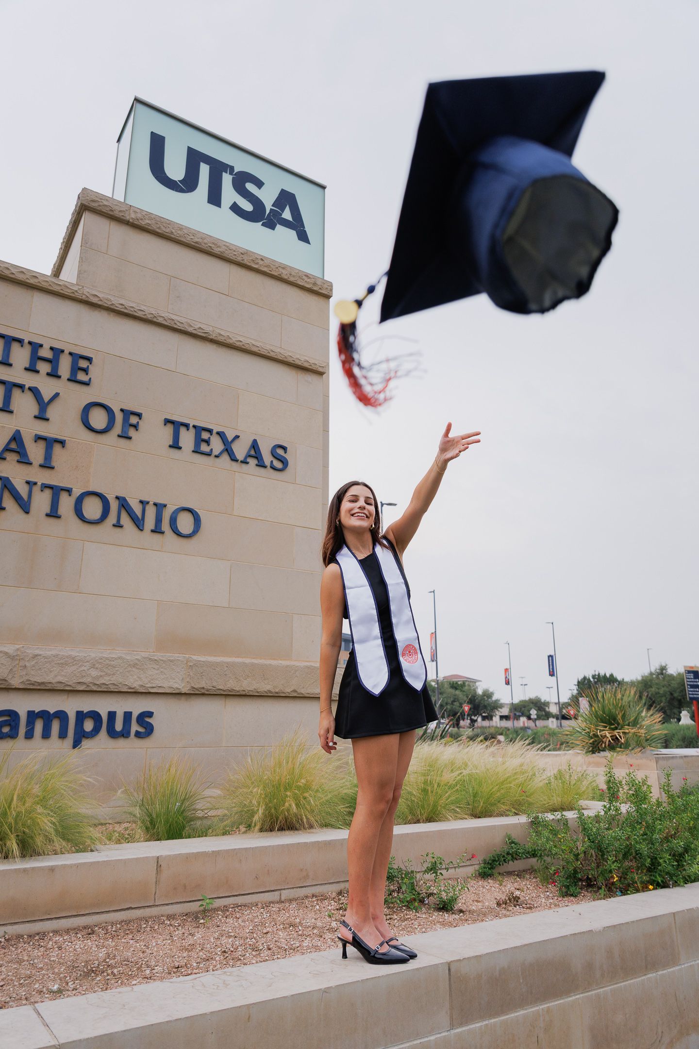 A UTSA graduate tosses her cap in the air in front of the University of Texas at San Antonio campus sign.