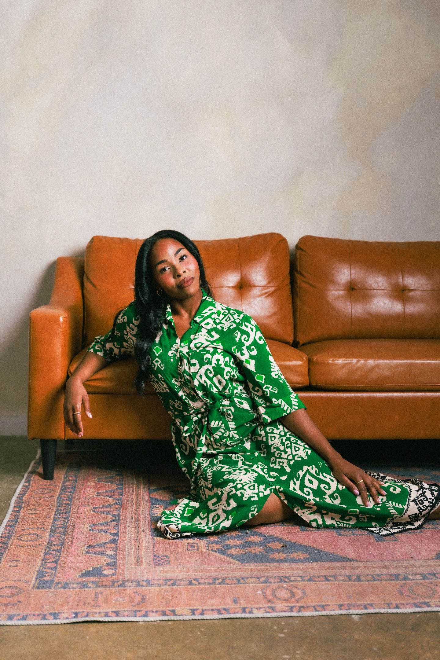 Woman in green floral dress sitting on floor leaning against brown leather sofa on patterned rug.