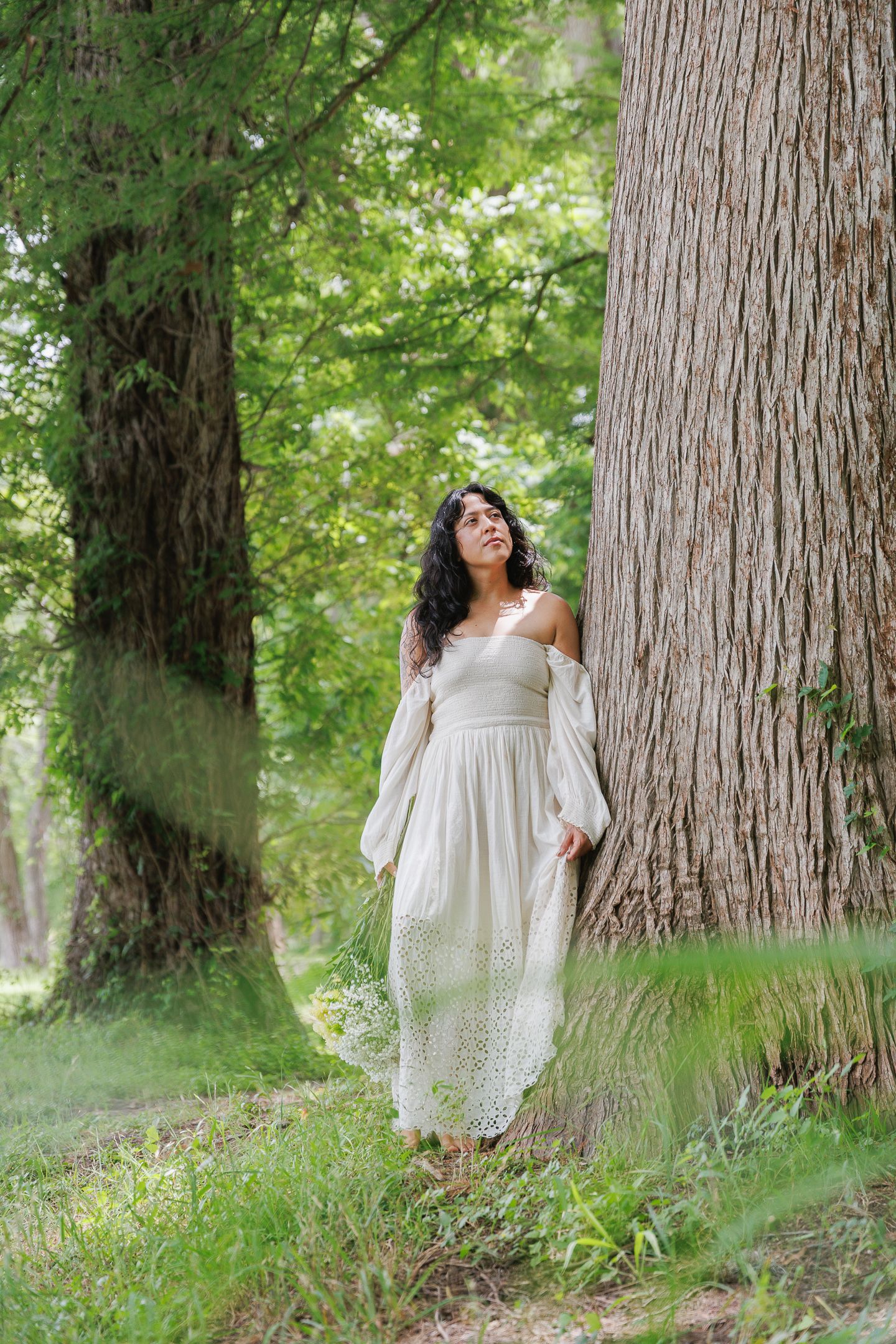Woman in white off-shoulder dress standing by large tree trunk in lush green forest.
