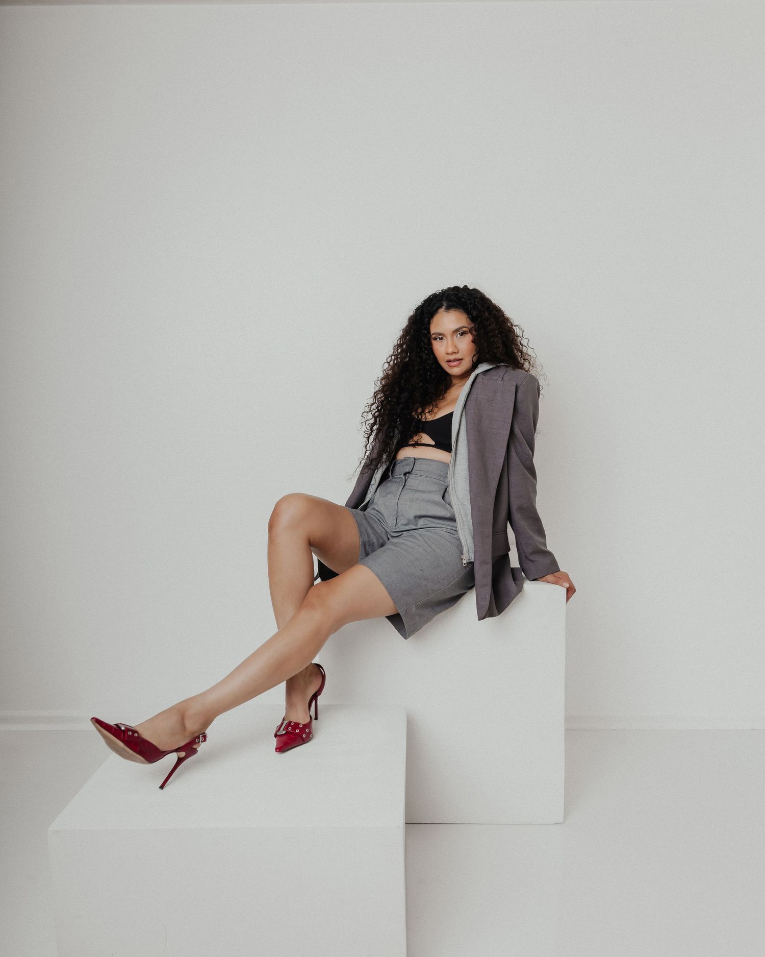 A fashion model poses on white geometric blocks wearing a grey blazer and red heels.