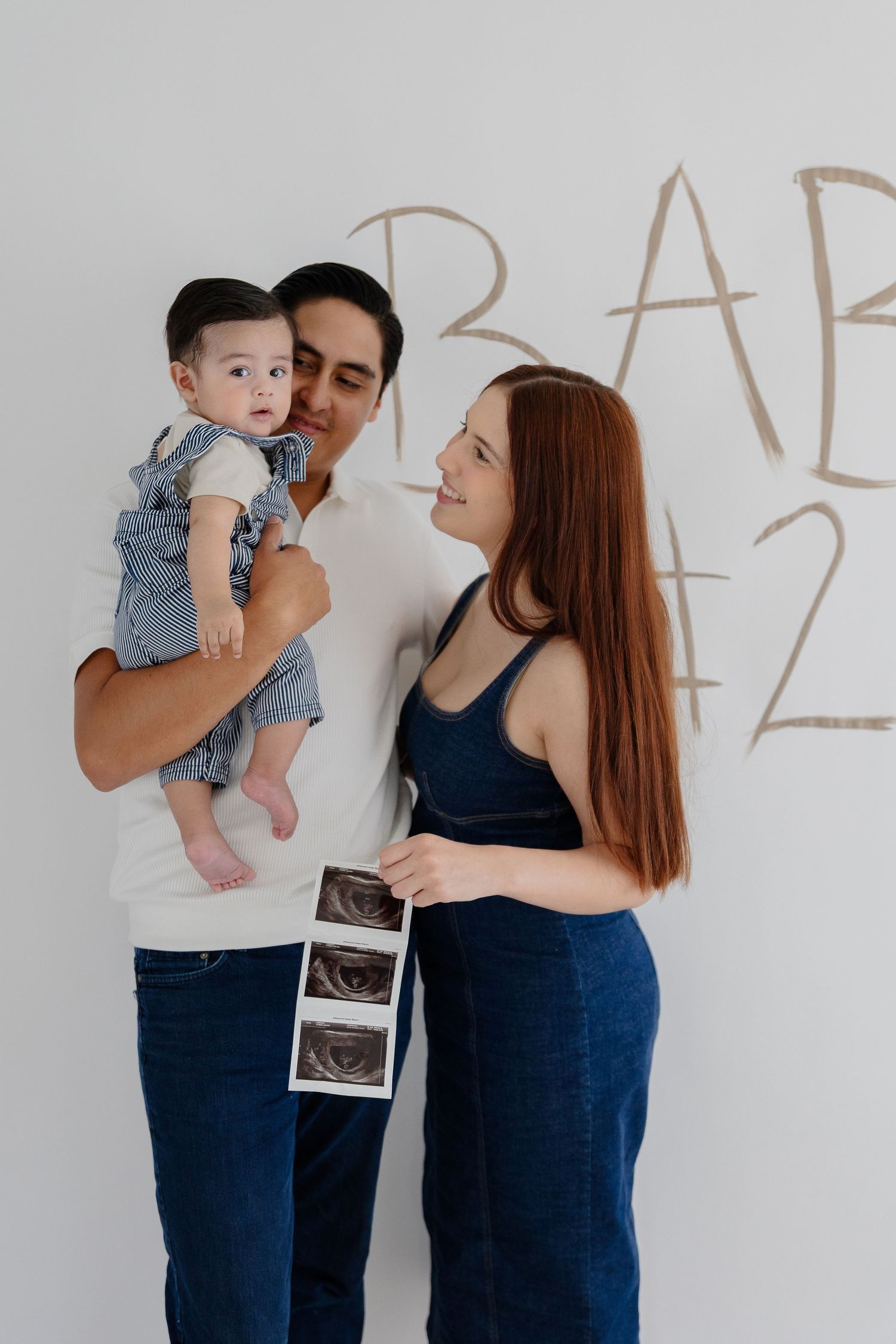 A family stands against a white wall holding ultrasound images during a pregnancy photoshoot.