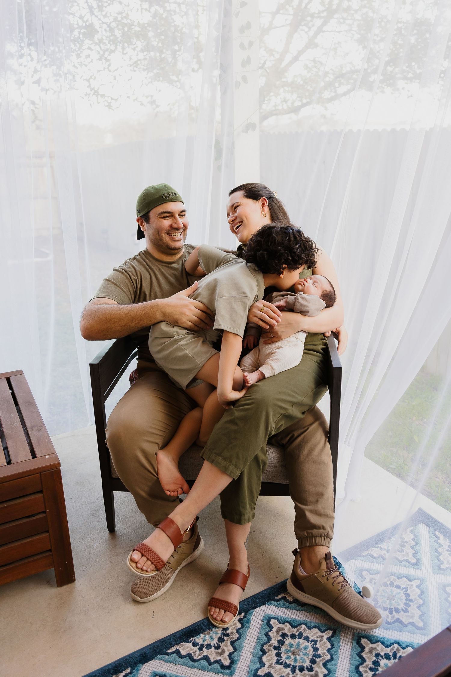 Family shares joyful laughter while sitting together on a chair in a bright room with sheer curtains and patterned rug.