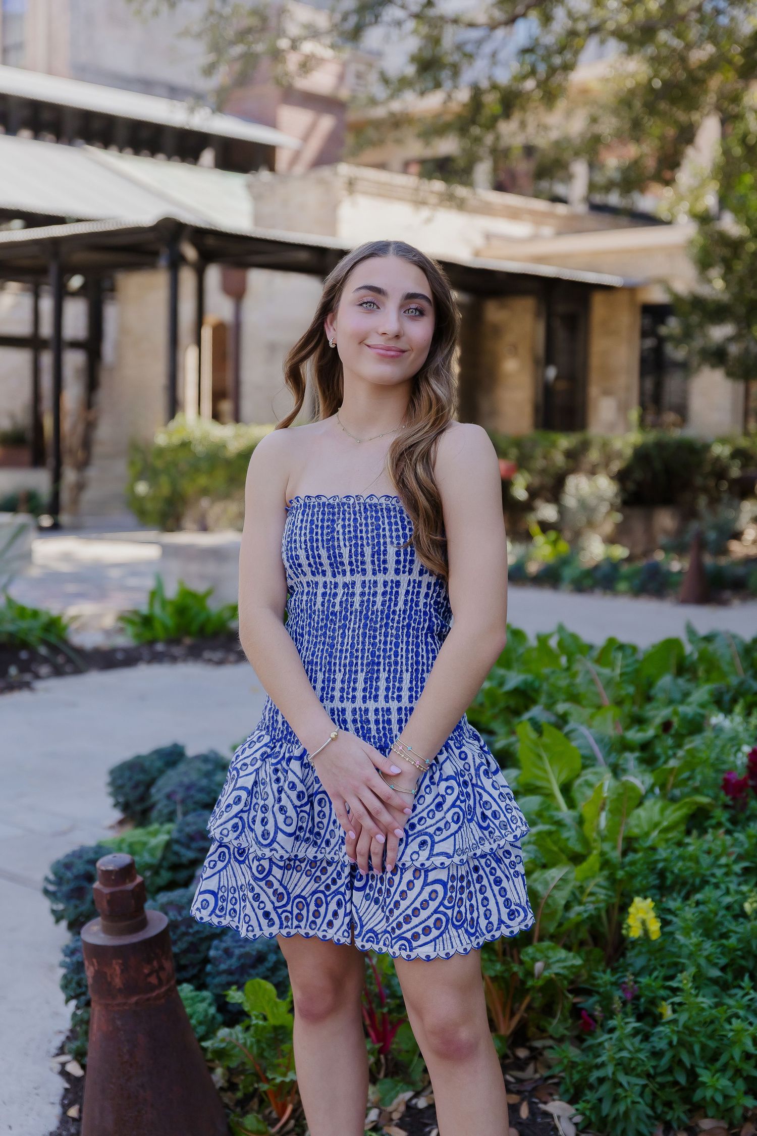 A person in a blue and white patterned sundress poses in front of a garden with flowering plants and greenery.