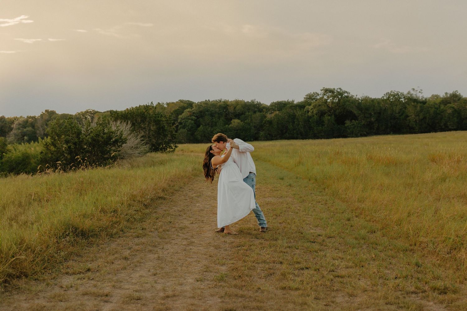 Romantic couple dancing together on a dirt path through a golden field at sunset.