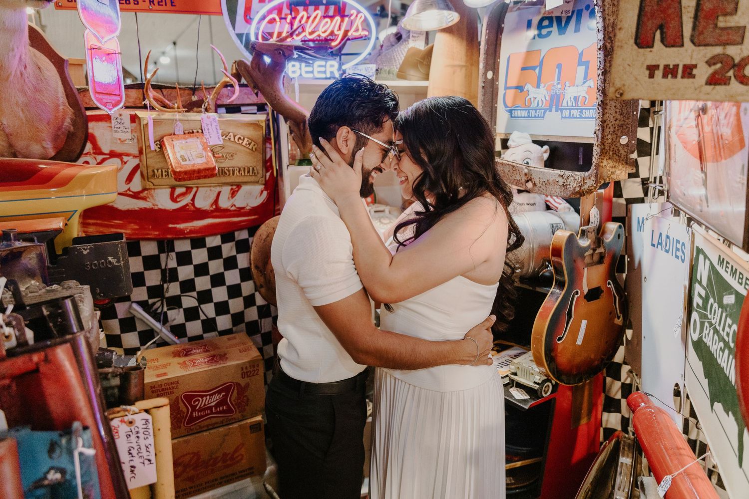 A romantic couple embraces in a vintage store surrounded by neon signs and nostalgic decor.