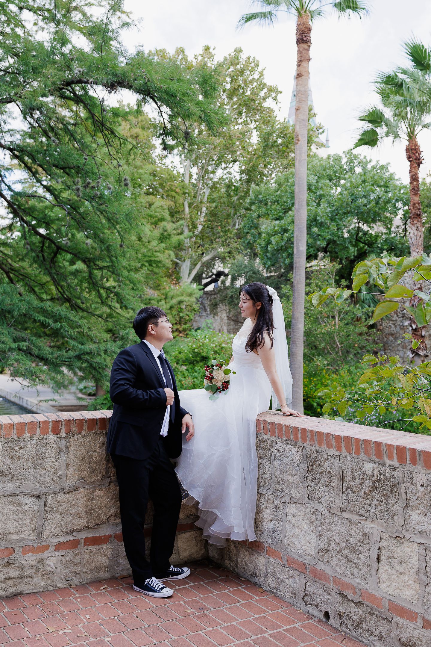 A couple in formal wedding attire shares a quiet moment on a stone terrace with palm trees and greenery in the background.