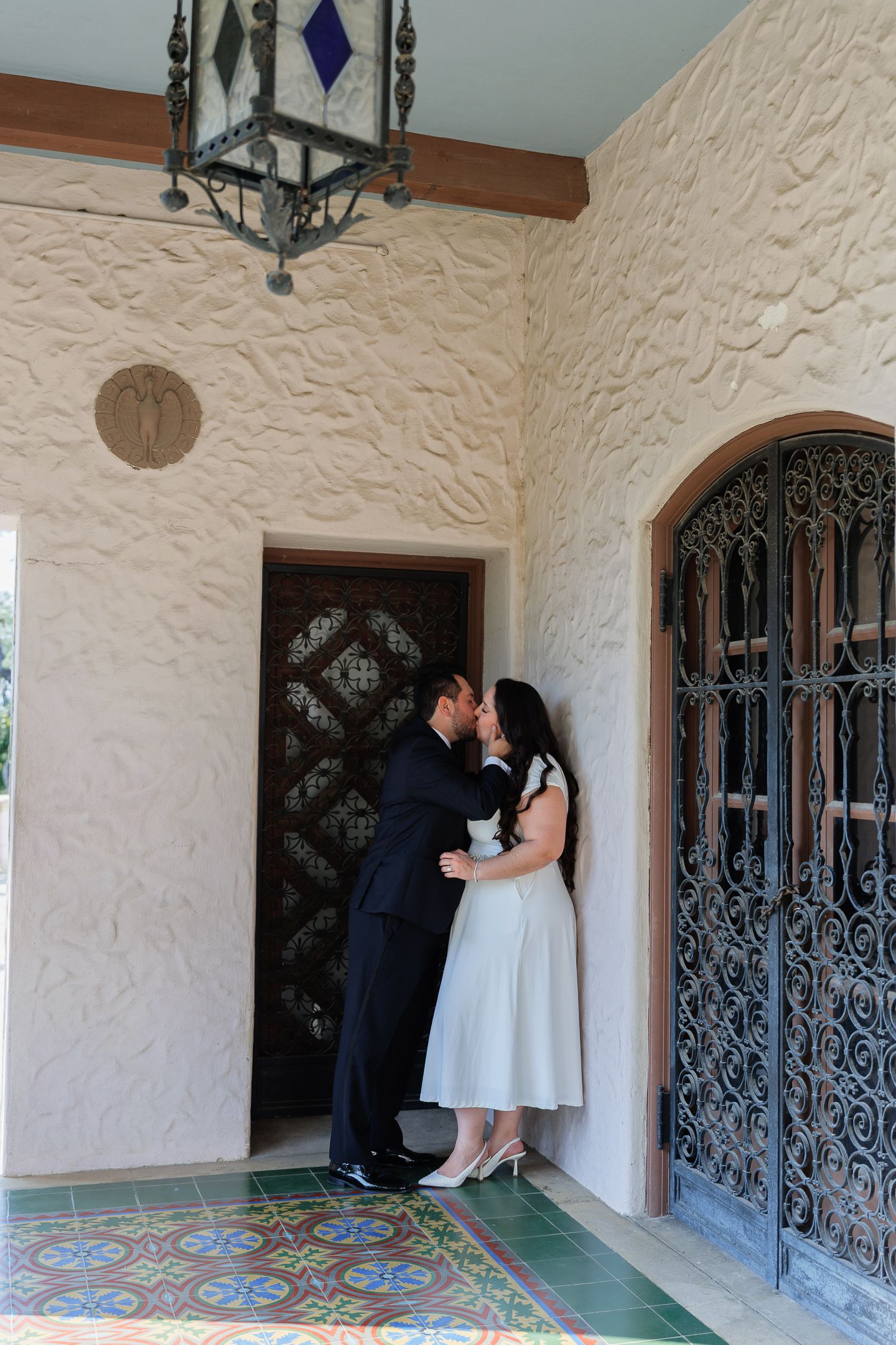 A couple shares a romantic moment in an elegant archway with ornate iron gates and decorative tiled floors.