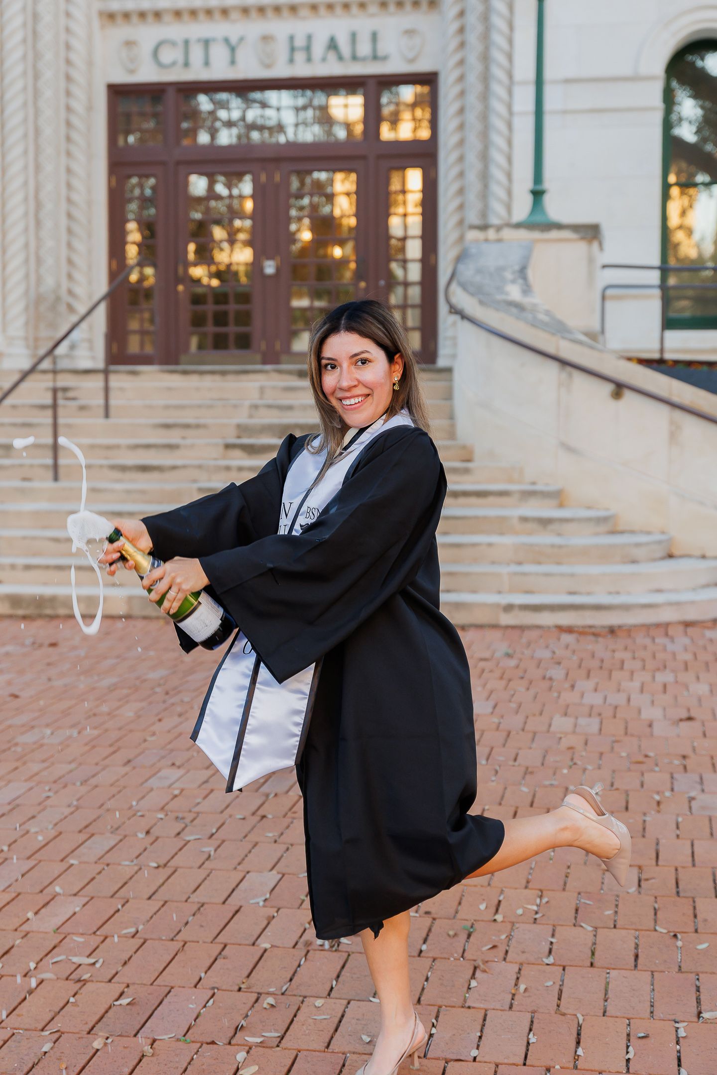 A graduate in a black gown poses playfully in front of a city hall building with brick pavement.