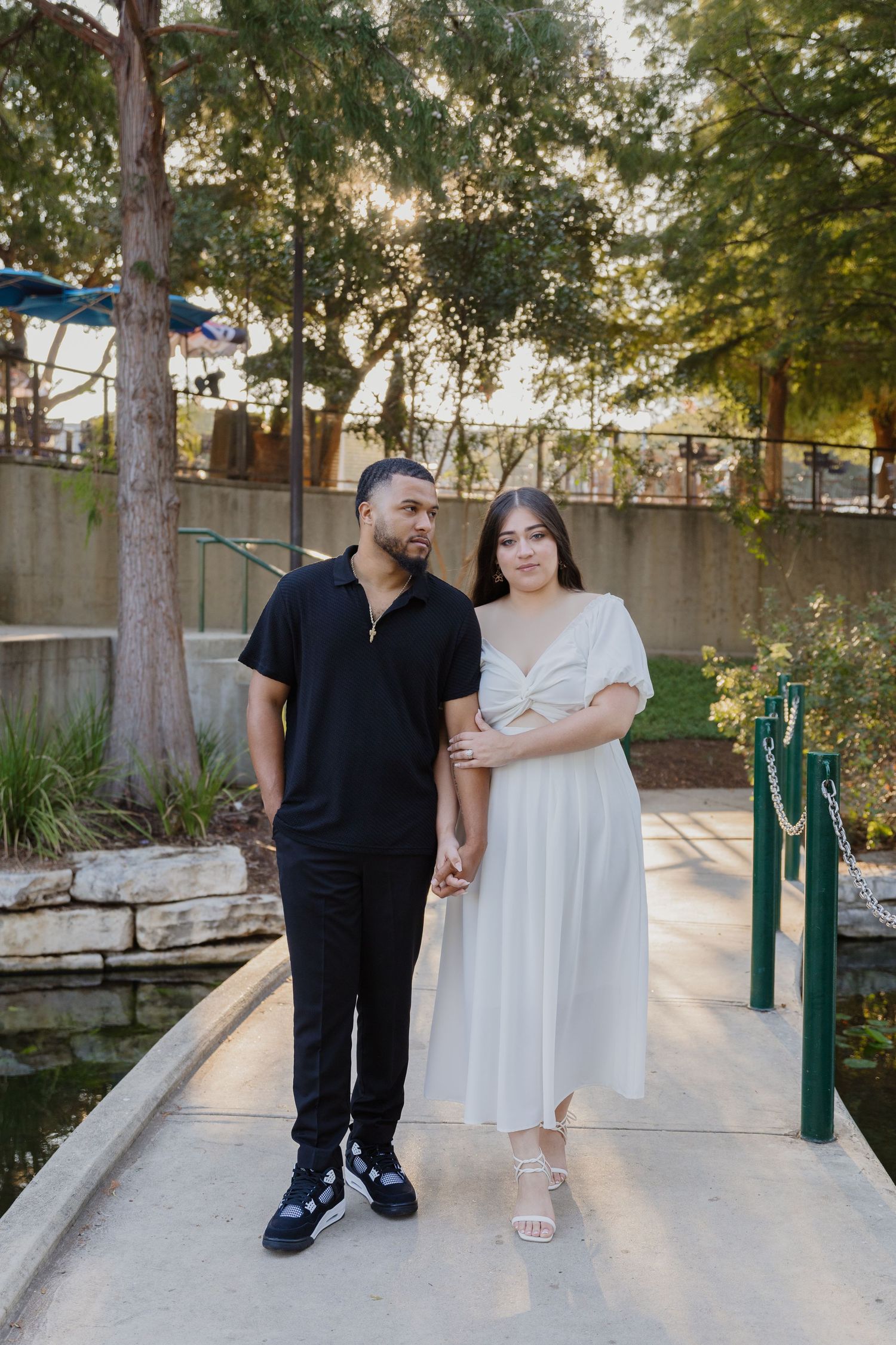 A couple poses together on a garden path surrounded by trees and landscaping.