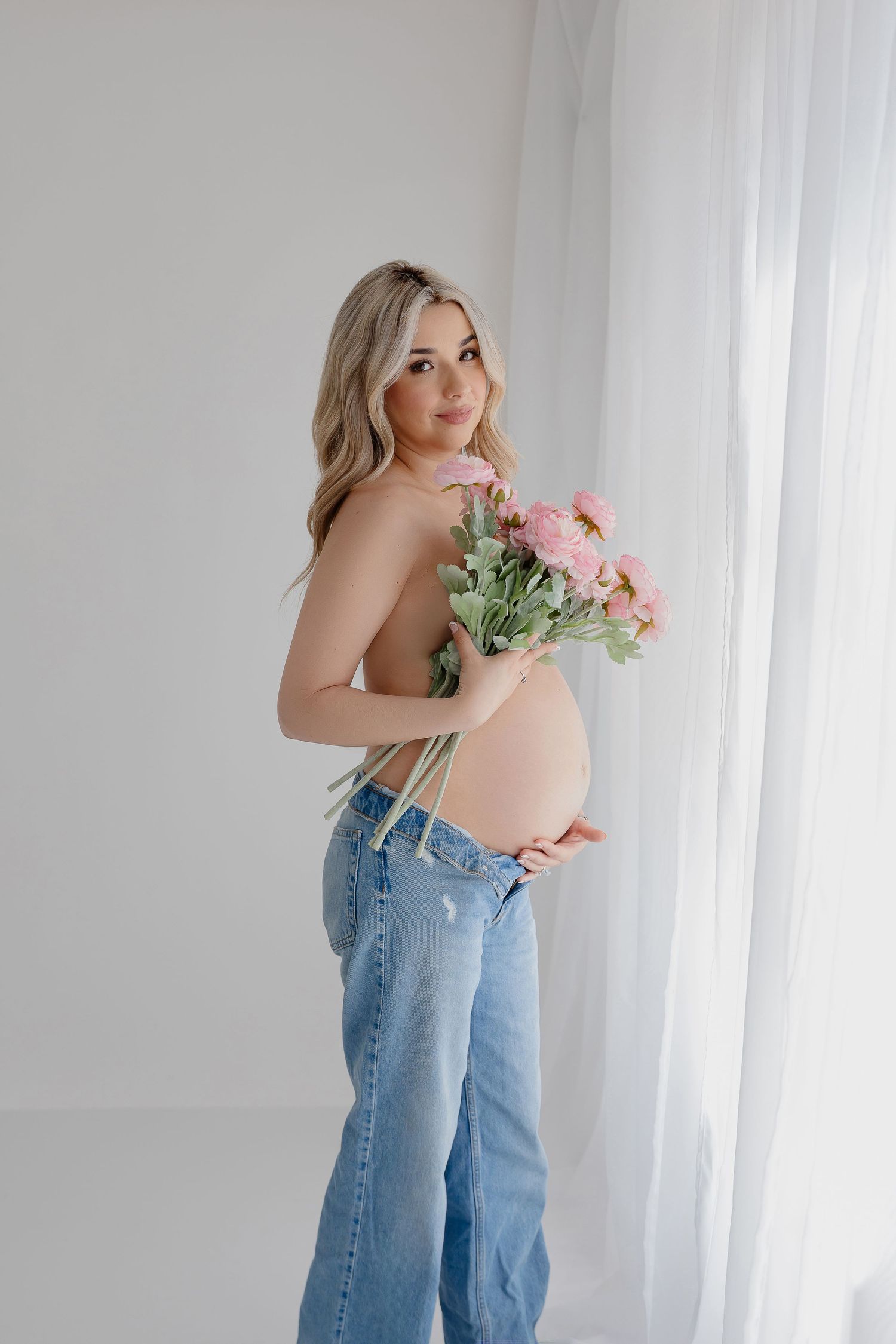 Expectant mother in jeans holding pink flowers during pregnancy photoshoot.