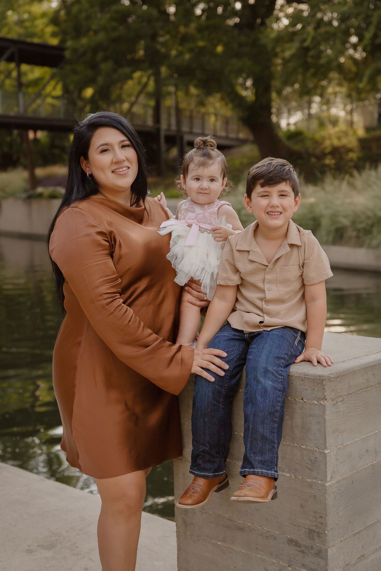 A family enjoys a casual autumn photoshoot by a pond with trees and a bridge in the background.