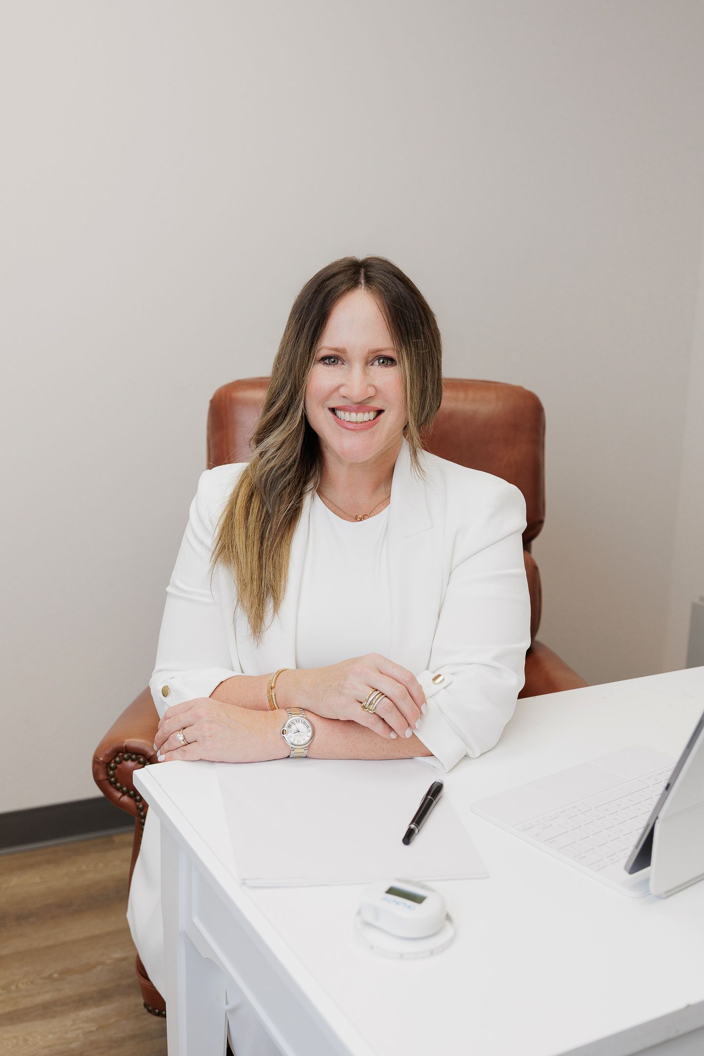 Professional wearing a white shirt sits at a desk in an office setting with a warm, welcoming expression.