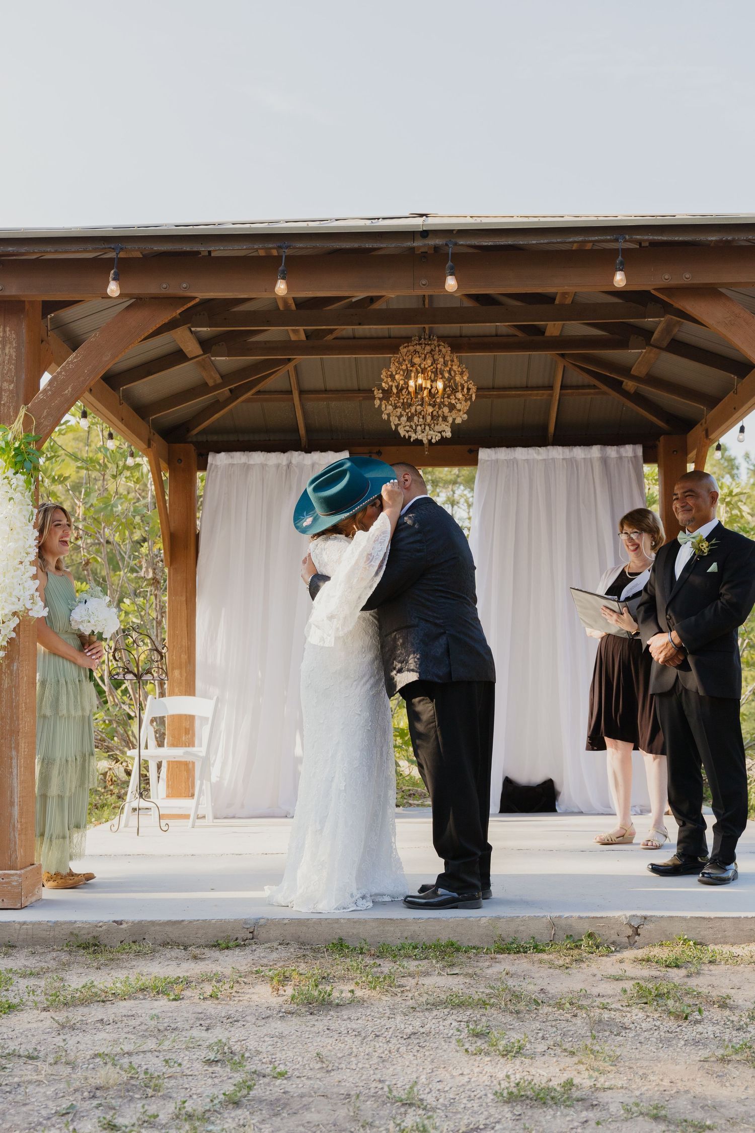 Wedding couple shares first kiss under rustic wooden pavilion with chandelier.