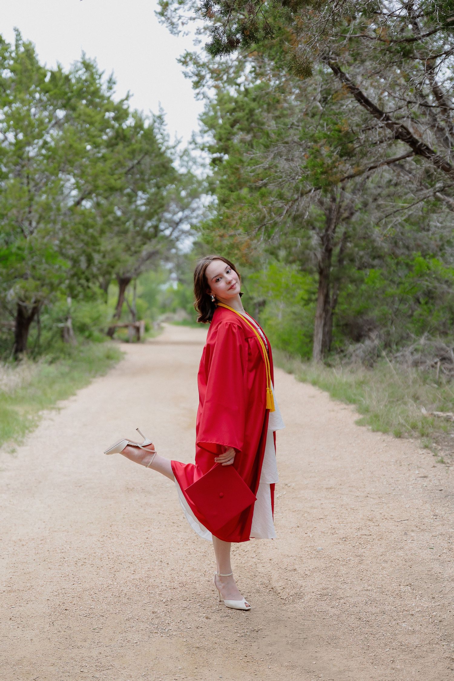 Graduate in red cap and gown playfully posing on nature trail.