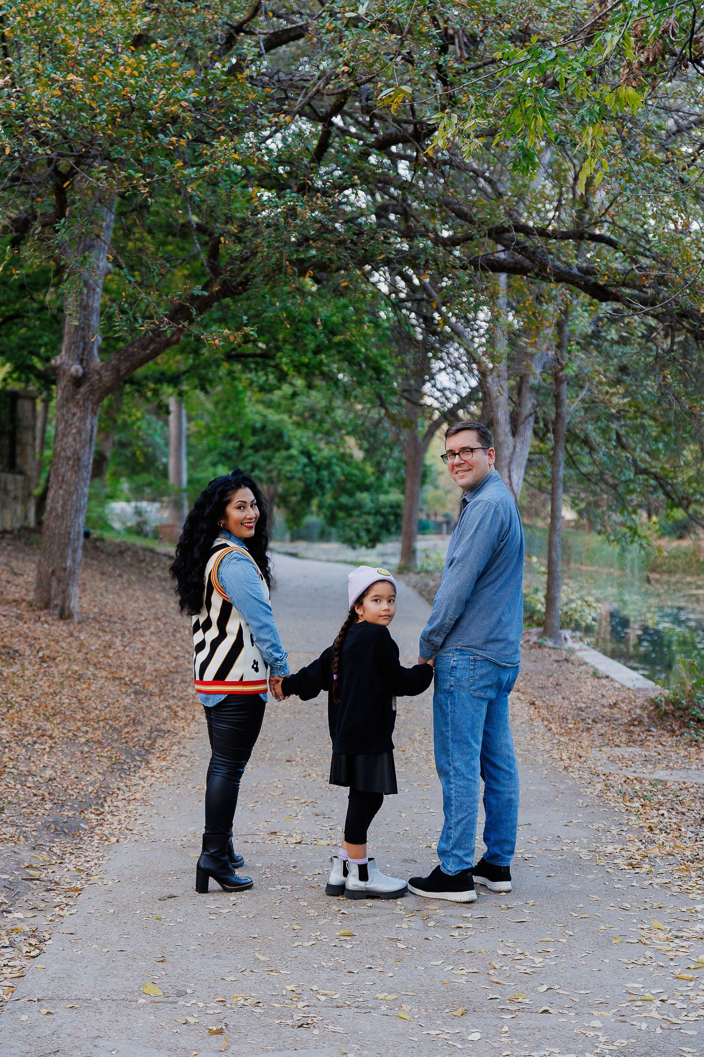 Three people walk together on a leaf-covered path in an autumn park setting.