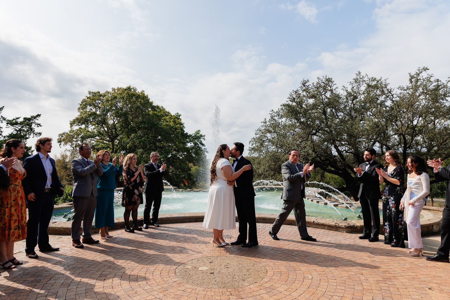 A couple embraces during their outdoor wedding ceremony on a brick path surrounded by guests and trees at sunset.
