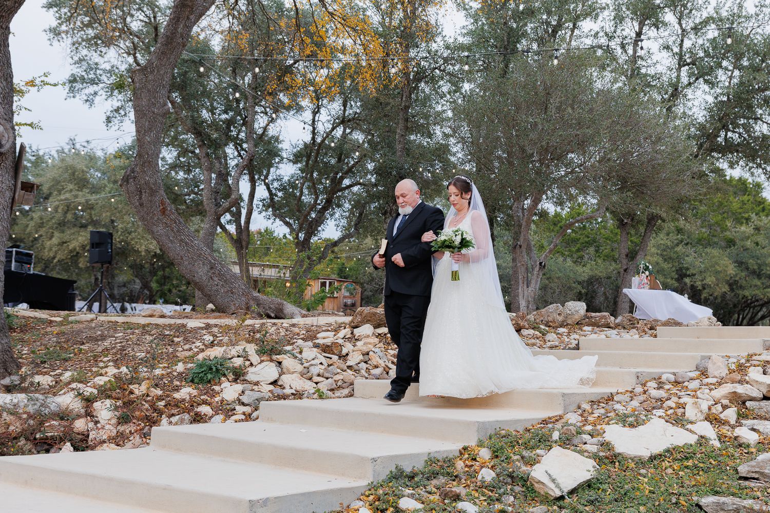 A couple in wedding attire walks along a winding stone pathway surrounded by autumn leaves in a scenic outdoor setting.