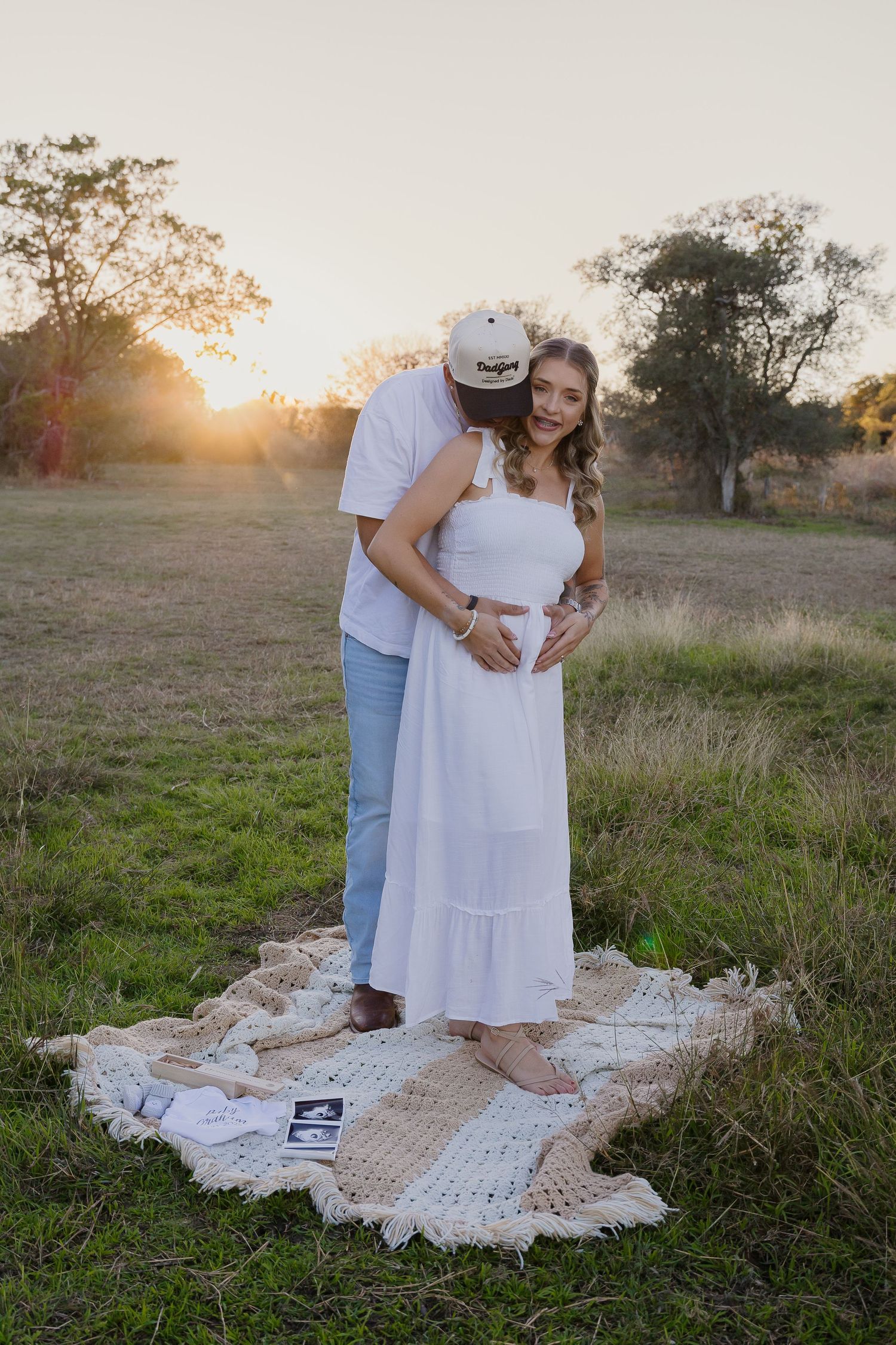 Sunset maternity photo of couple standing on vintage blanket with ultrasound picture.