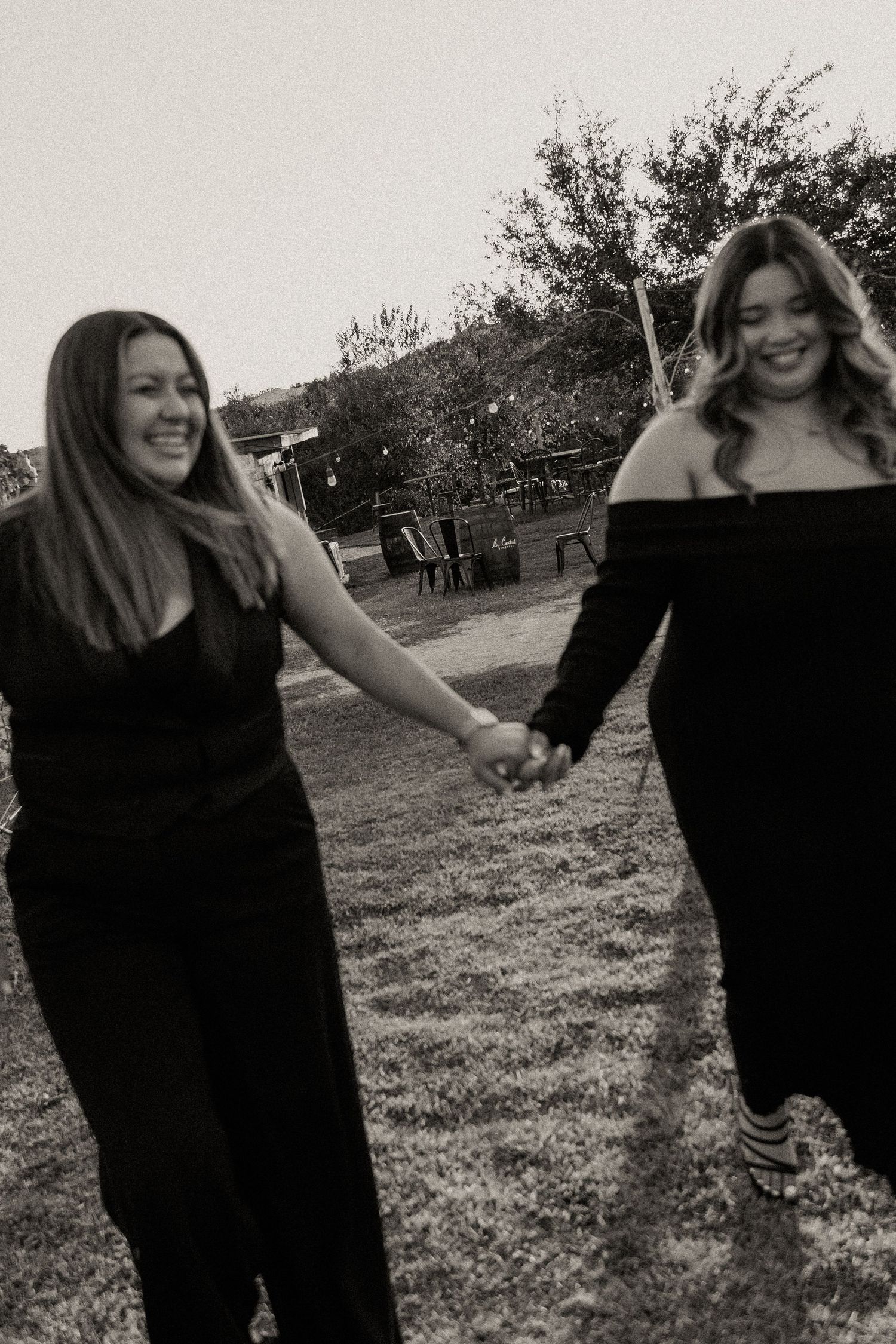 Two women in black outfits laugh while holding hands in outdoor garden setting.