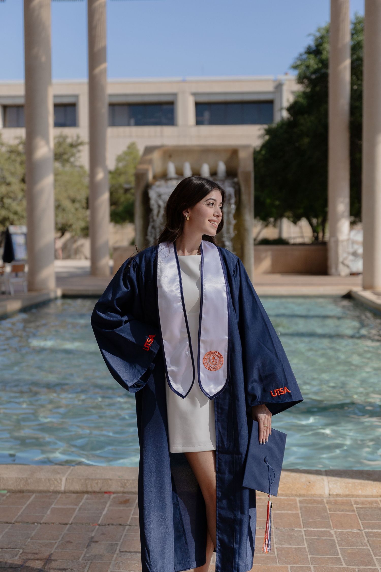 Graduate in navy blue graduation gown smiles while posing by campus fountain.