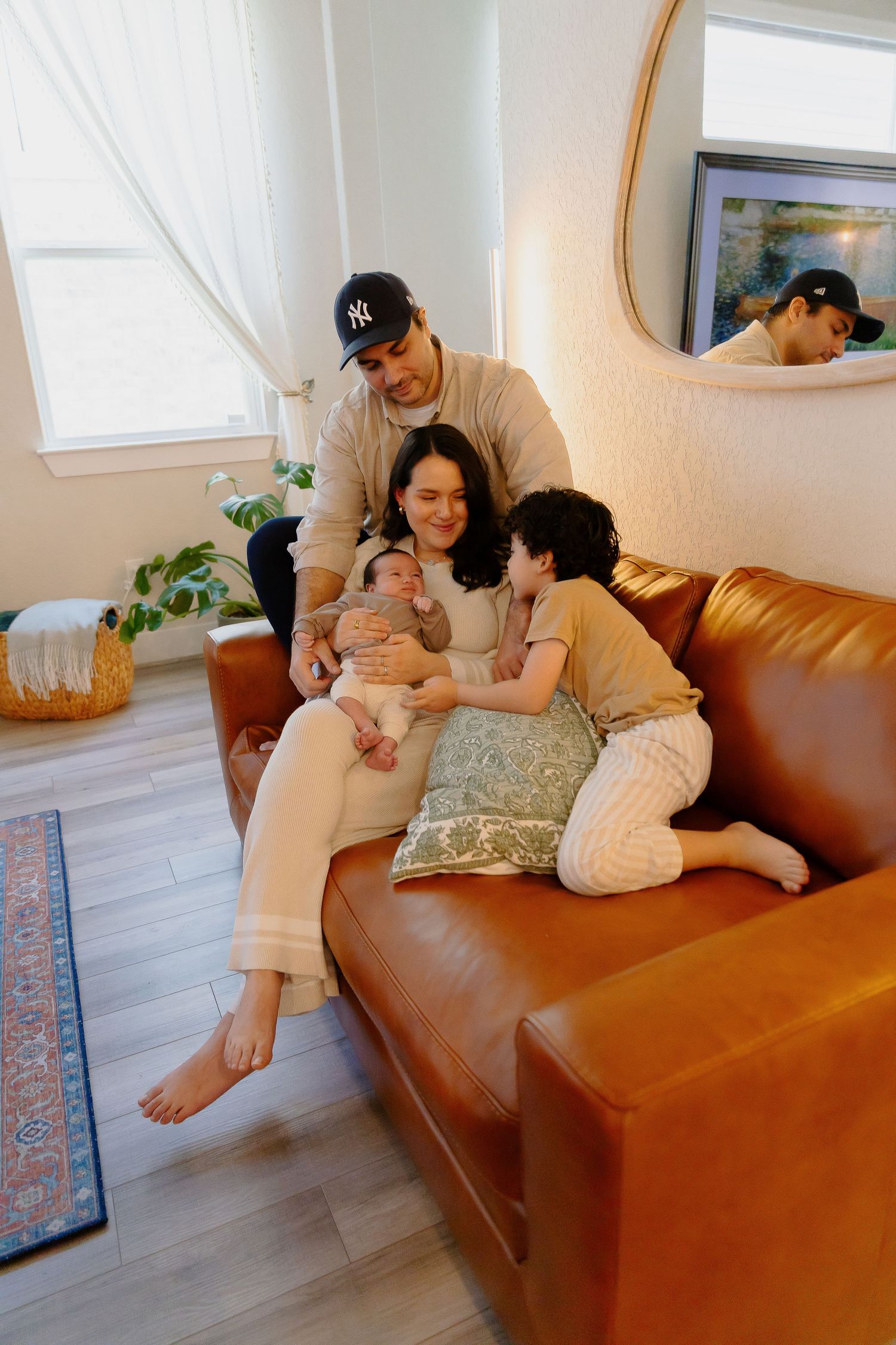 Family shares tender moment together on brown leather couch with infant and plants in background.