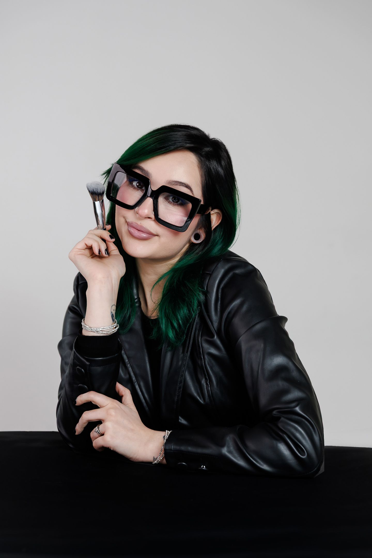 Woman with green-tipped hair and large glasses holds a makeup brush in a black leather jacket against a gray background.