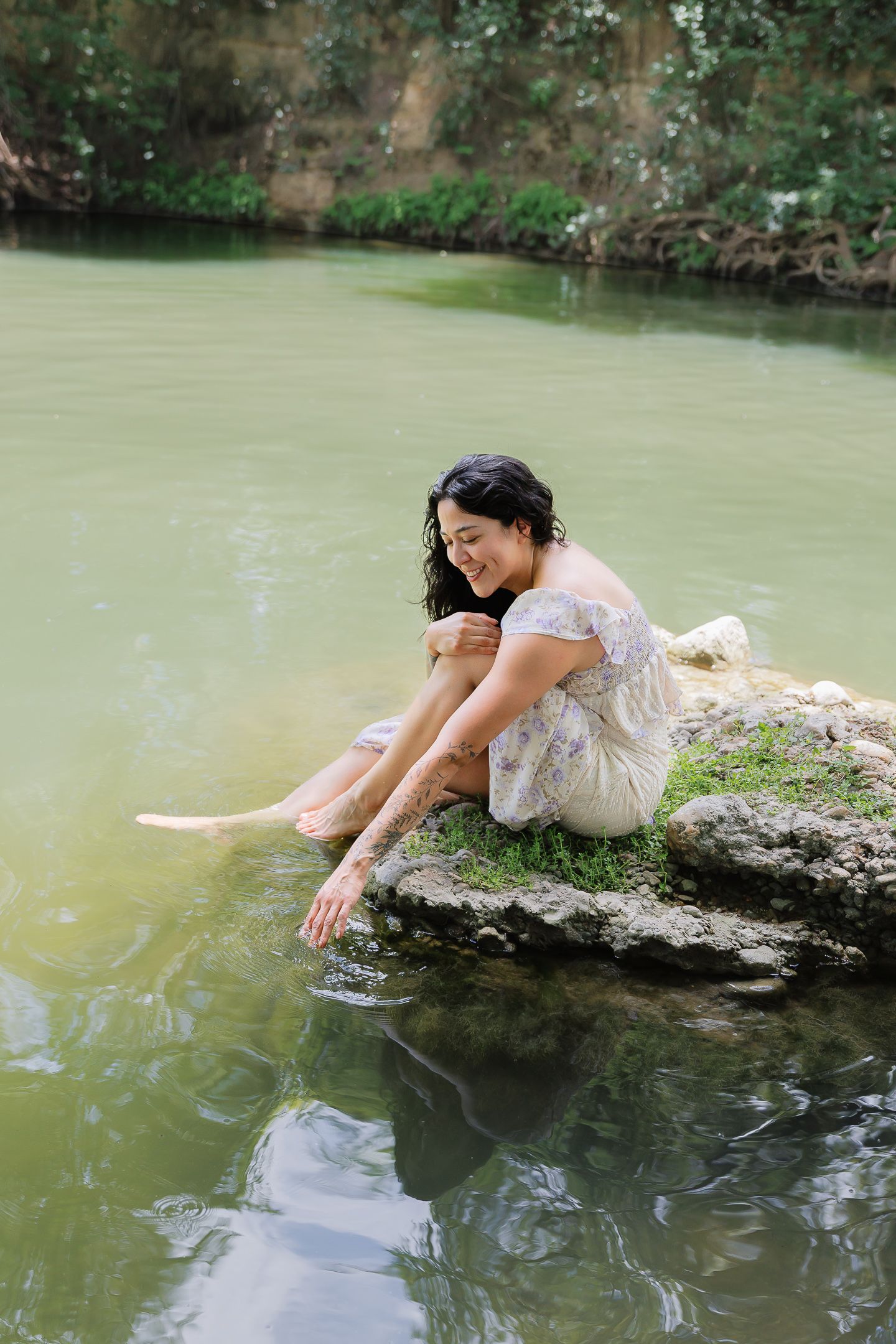 A smiling woman in a floral dress sits on a mossy rock, dipping her hand into a serene green river outdoors.