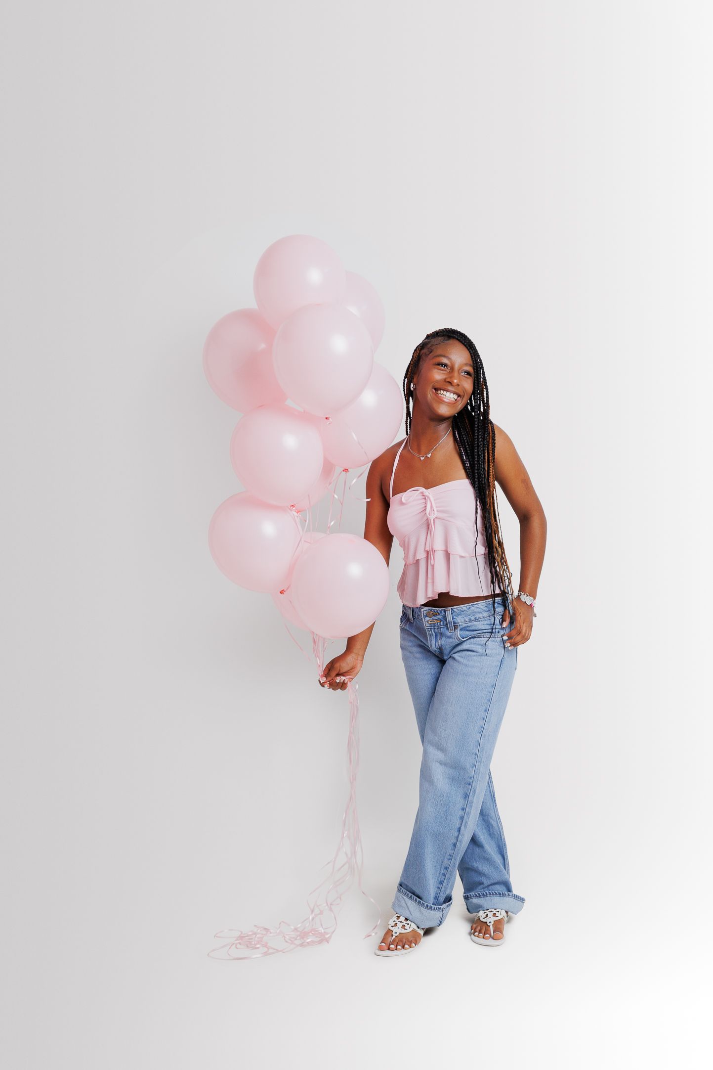 Smiling young woman with braids holding pink balloons, wearing a pink crop top and wide-leg jeans on white background.