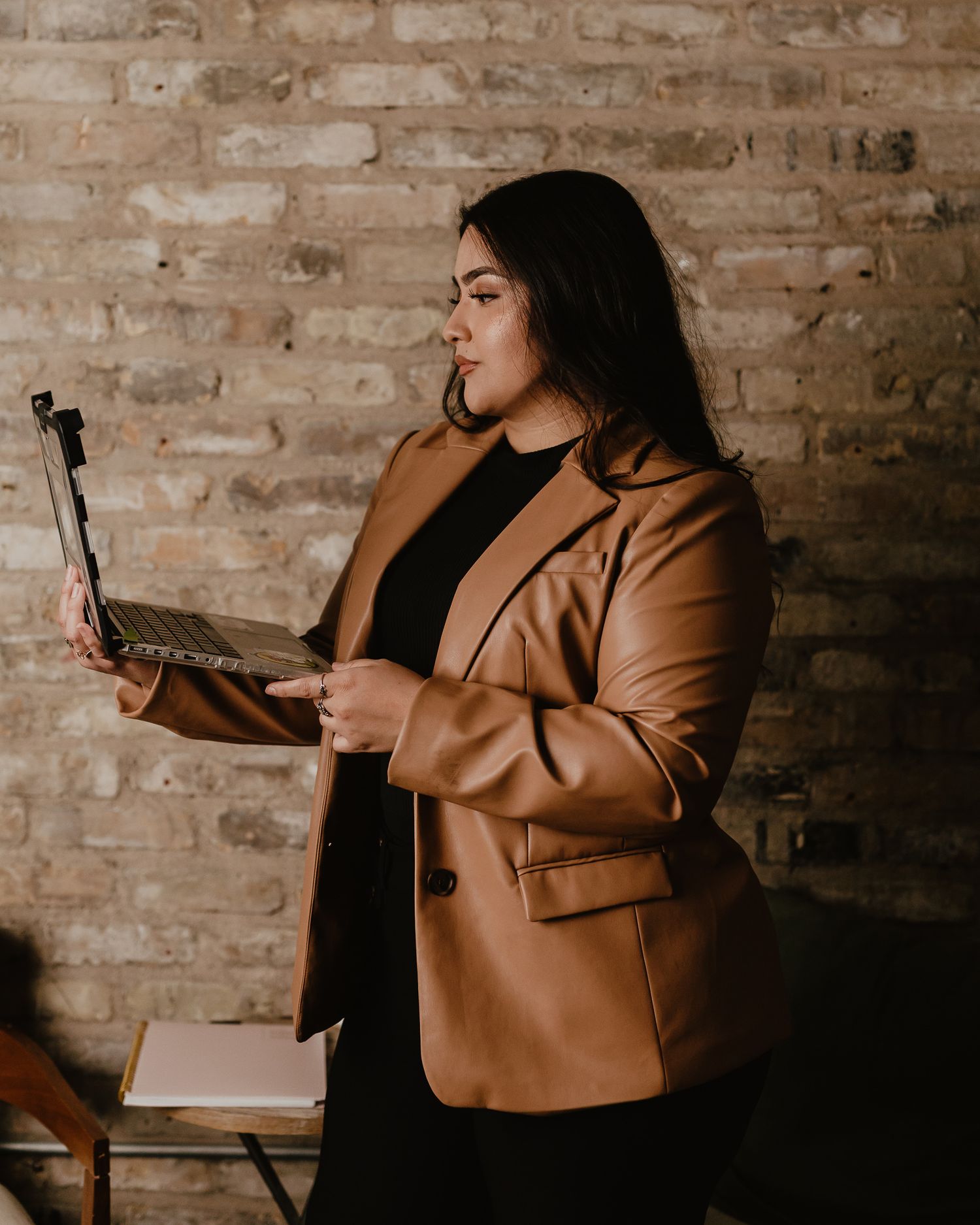 Professional in tan blazer presents or speaks in brick-walled meeting space.