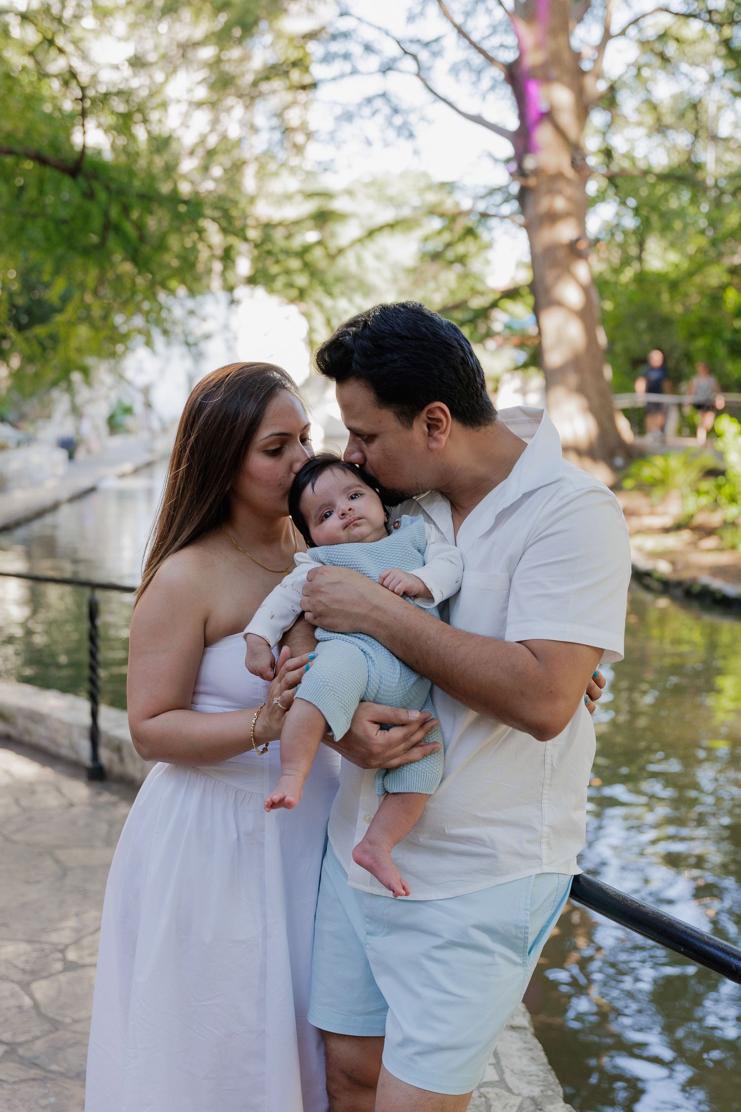 A couple in white outfits sharing a tender moment with their baby by a pond in a park setting.