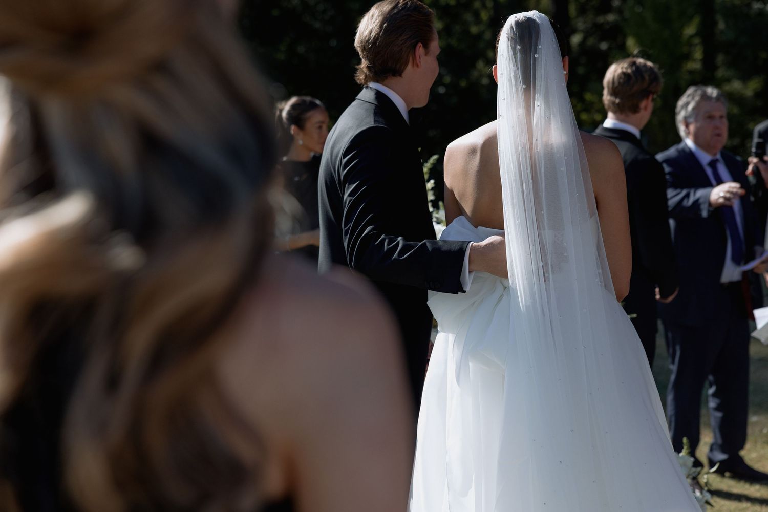 A bride in a white dress and veil stands among wedding guests during an outdoor celebration.