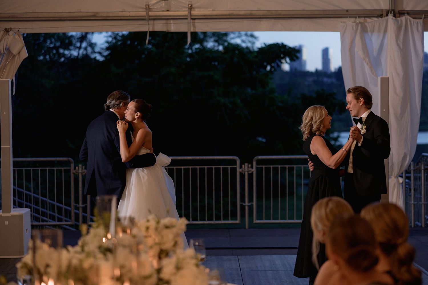 Wedding ceremony taking place at dusk under an illuminated tent with city skyline in background.