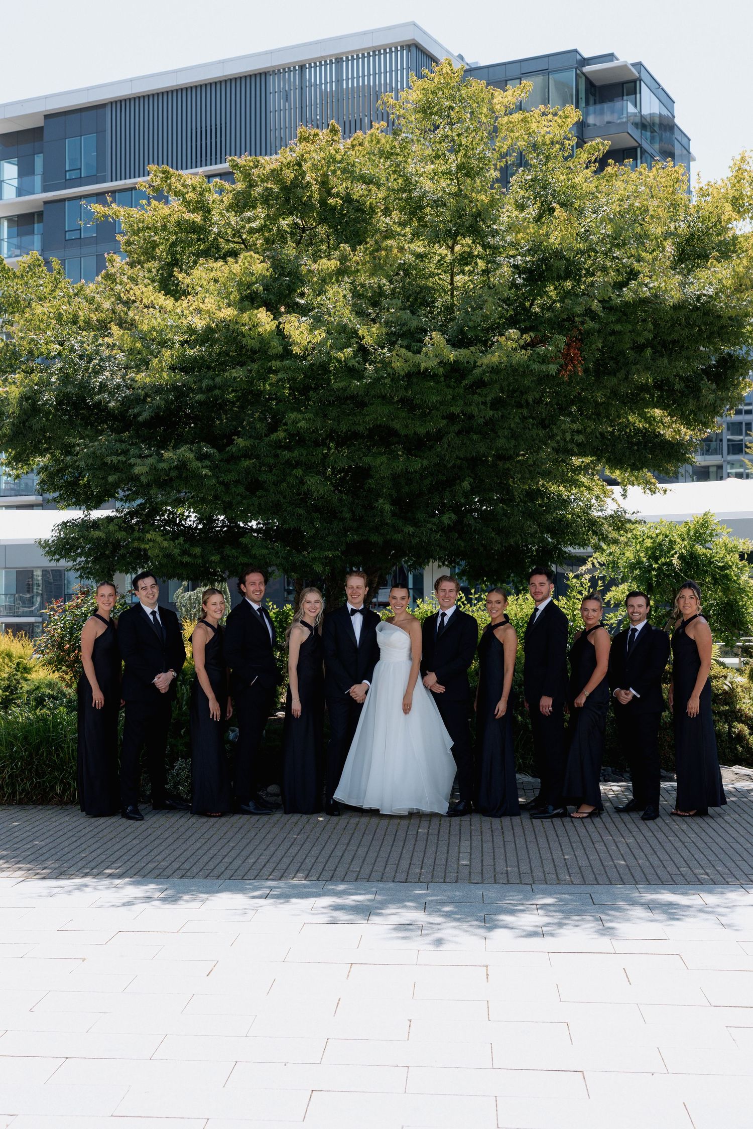 Formal wedding group photo with bridal party arranged in front of lush trees and glass buildings at Marriott Hotel in Vancouver