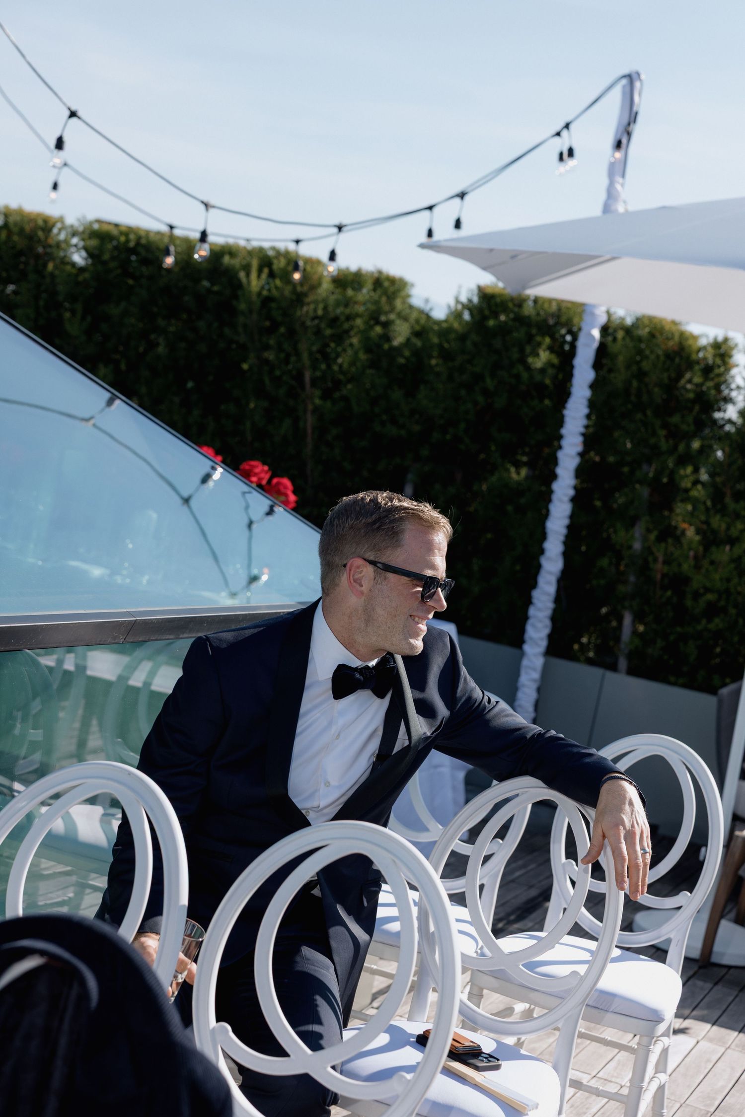 White chairs arranged for an outdoor ceremony with string lights overhead and hedges in the background.
