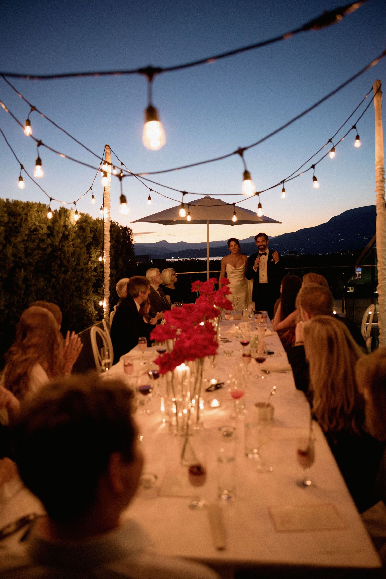 Evening wedding reception under string lights with mountain views and red floral centerpieces.