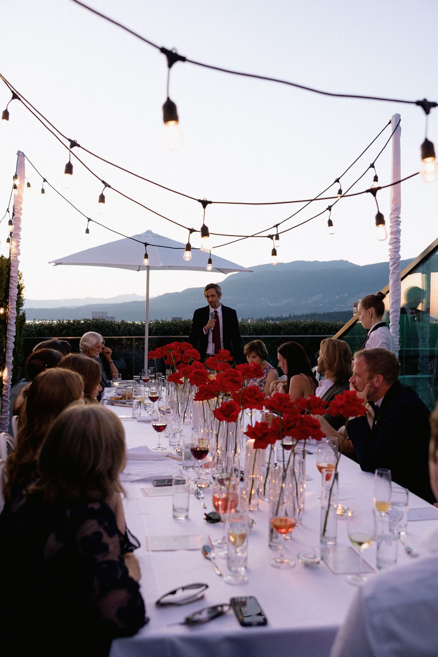 An outdoor wedding reception dinner table with red flowers and string lights against a mountain backdrop at dusk.