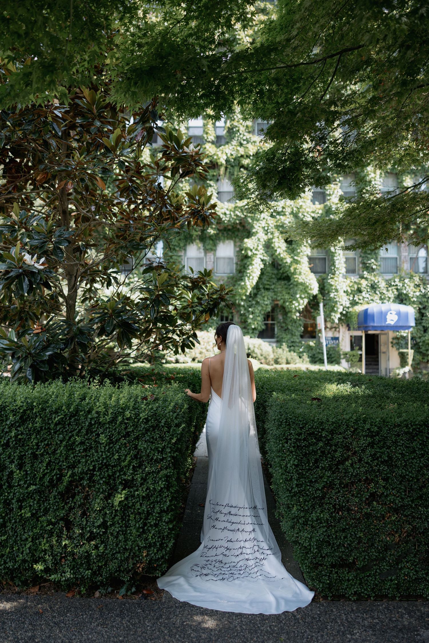 A flowing white wedding dress with intricate details photographed against a lush green hedge backdrop.