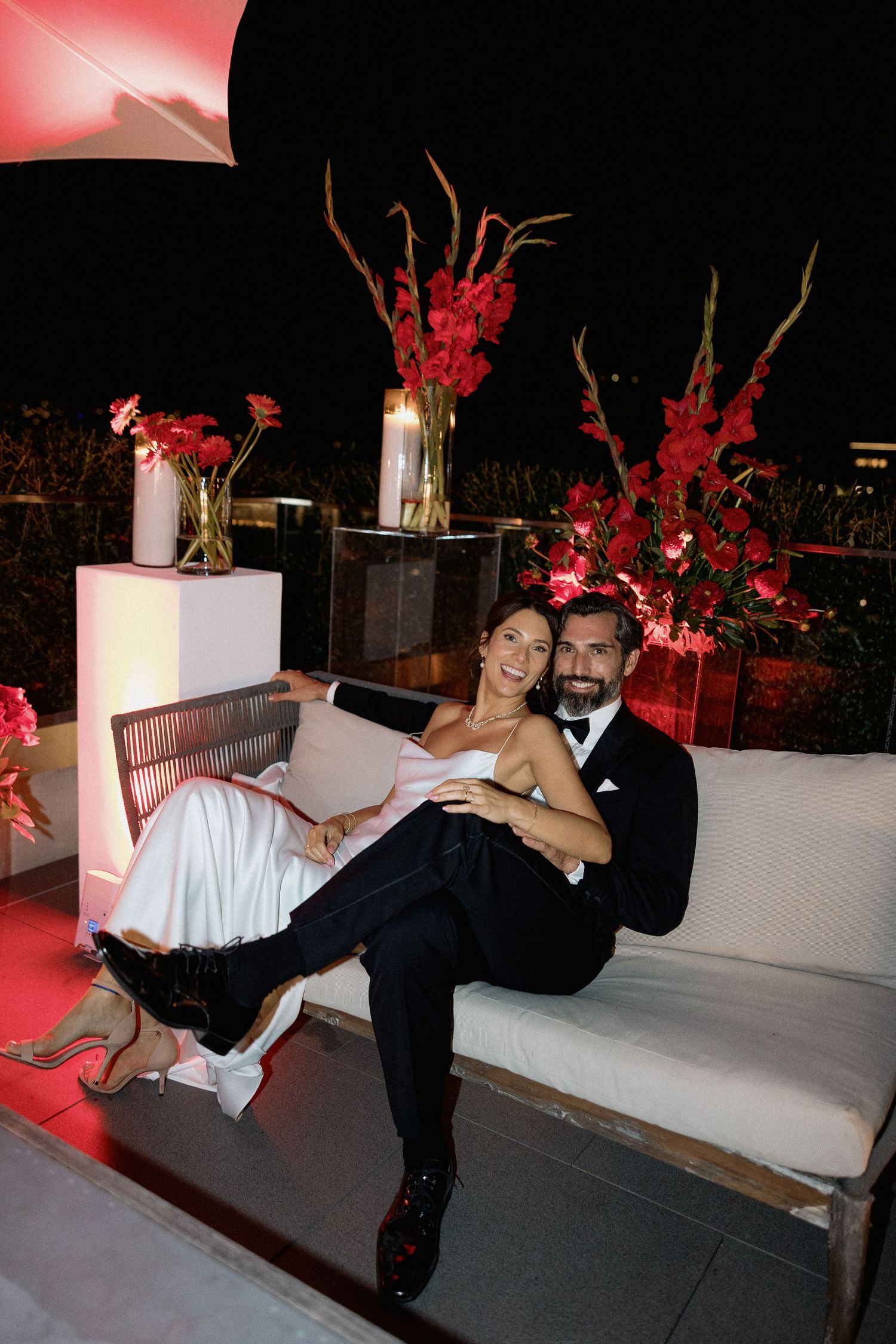 Couple in formal attire posing on outdoor terrace with red floral decorations at night in the night of Vancouver