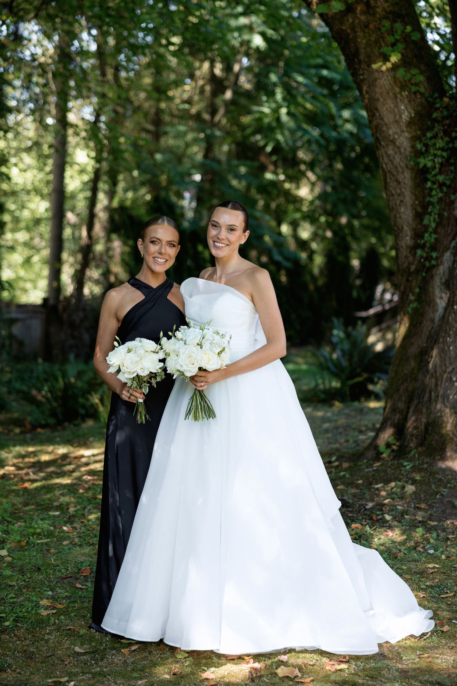 Wedding photo in a forest setting showing a bride in white gown and attendant in black dress holding white bouquets.