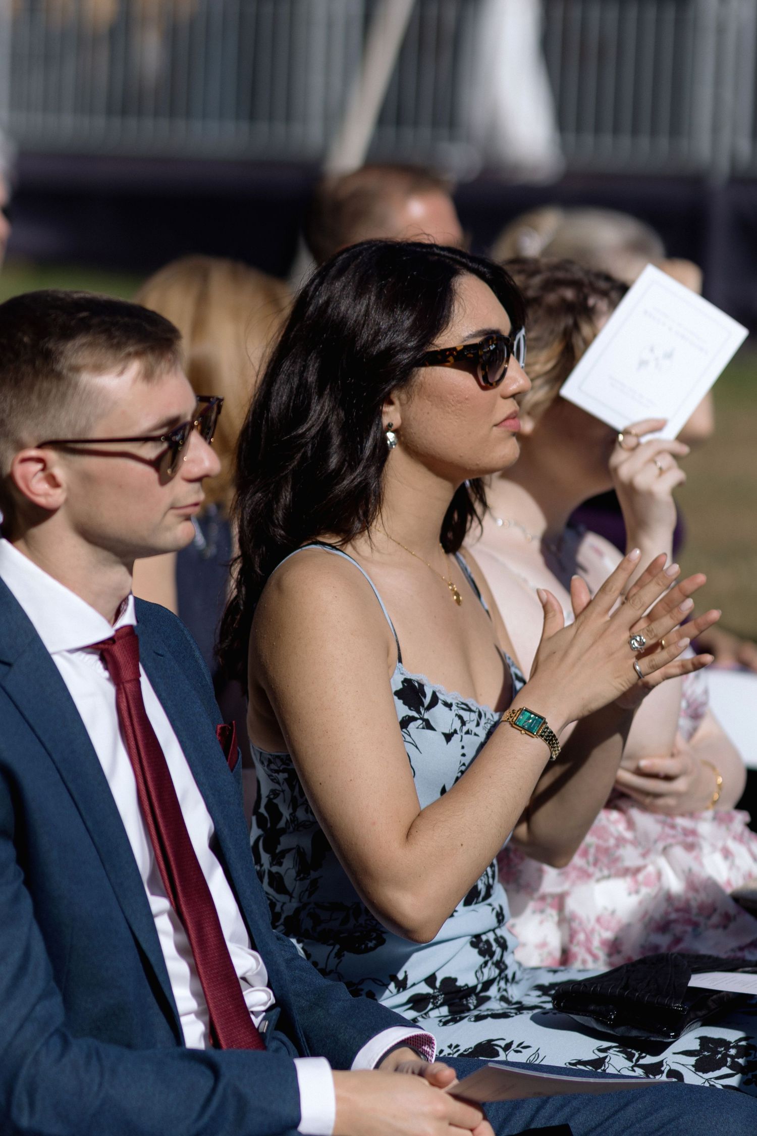 Wedding guests in formal attire holding ceremony programs during outdoor celebration.