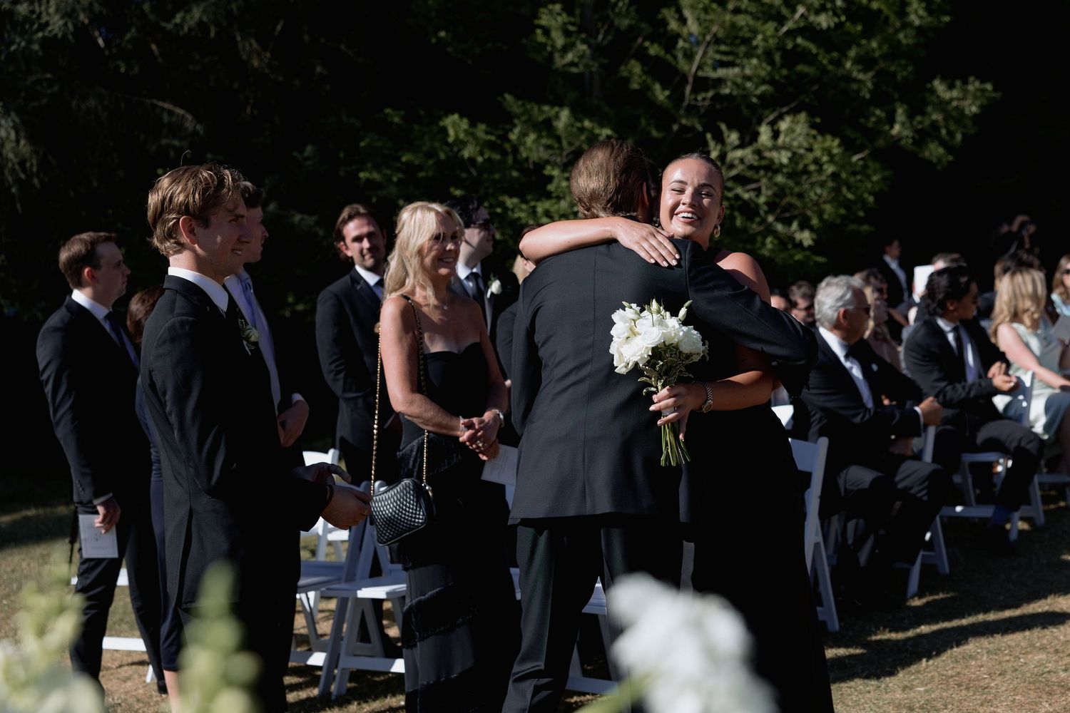 Wedding guests gather on a sunny day for an outdoor ceremony among trees.