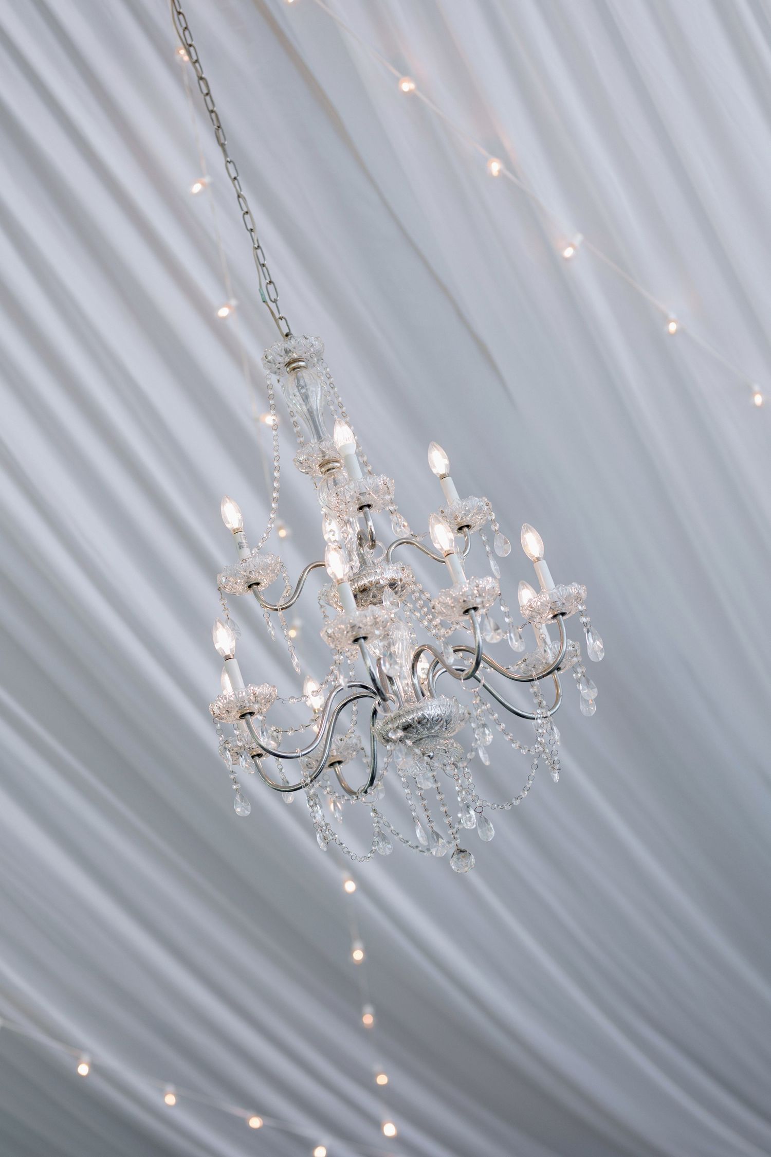 Crystal chandelier hanging from white draped ceiling with twinkling fairy lights.