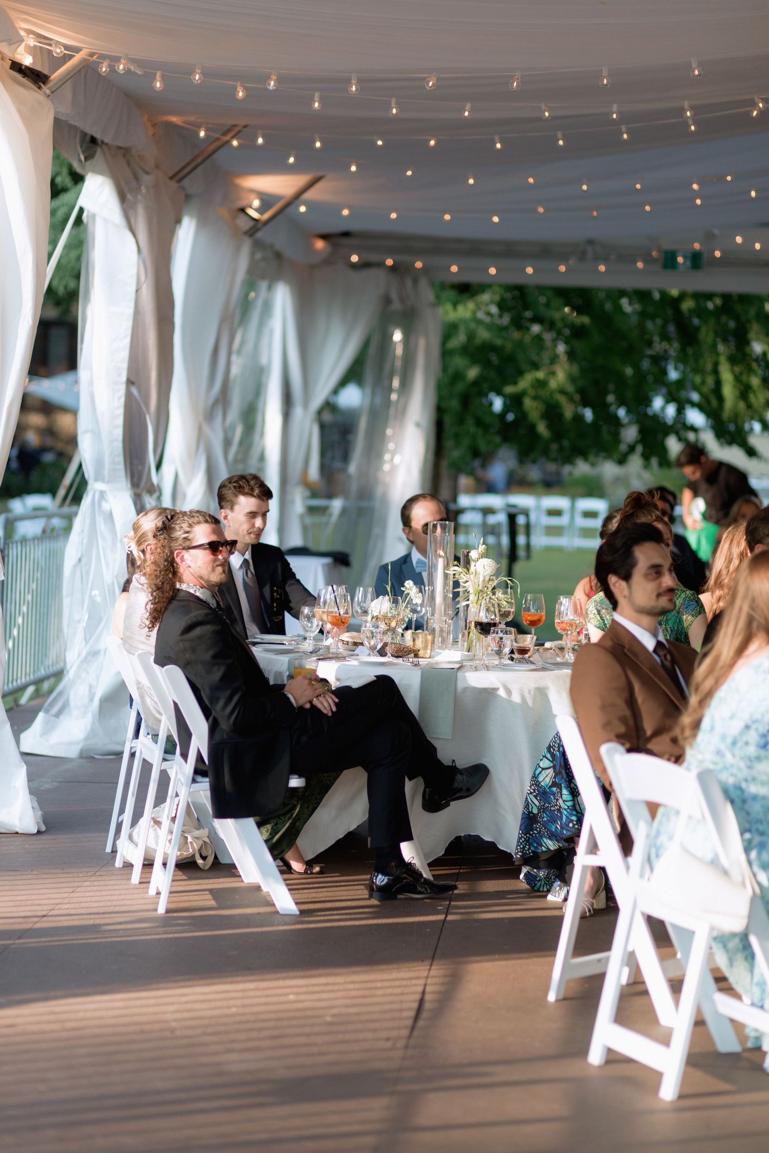Reception guests enjoying dinner under a white tent with twinkling lights and outdoor views.