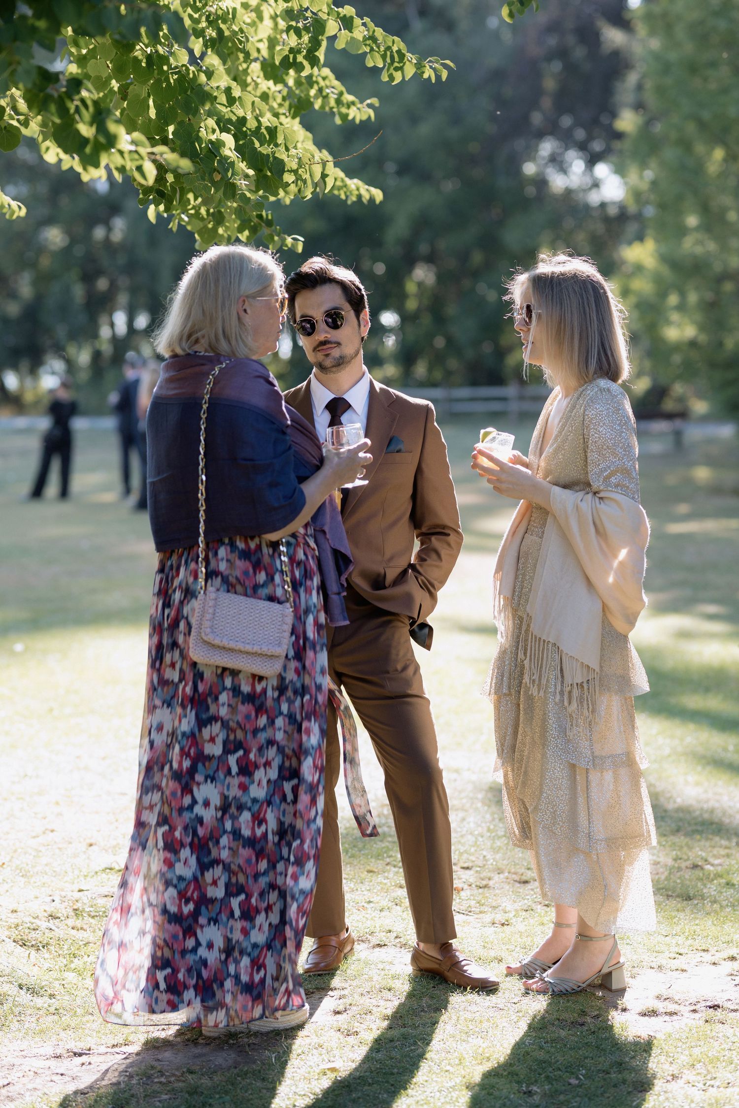 Wedding guests in floral dresses socialize on a sunlit lawn during the reception.