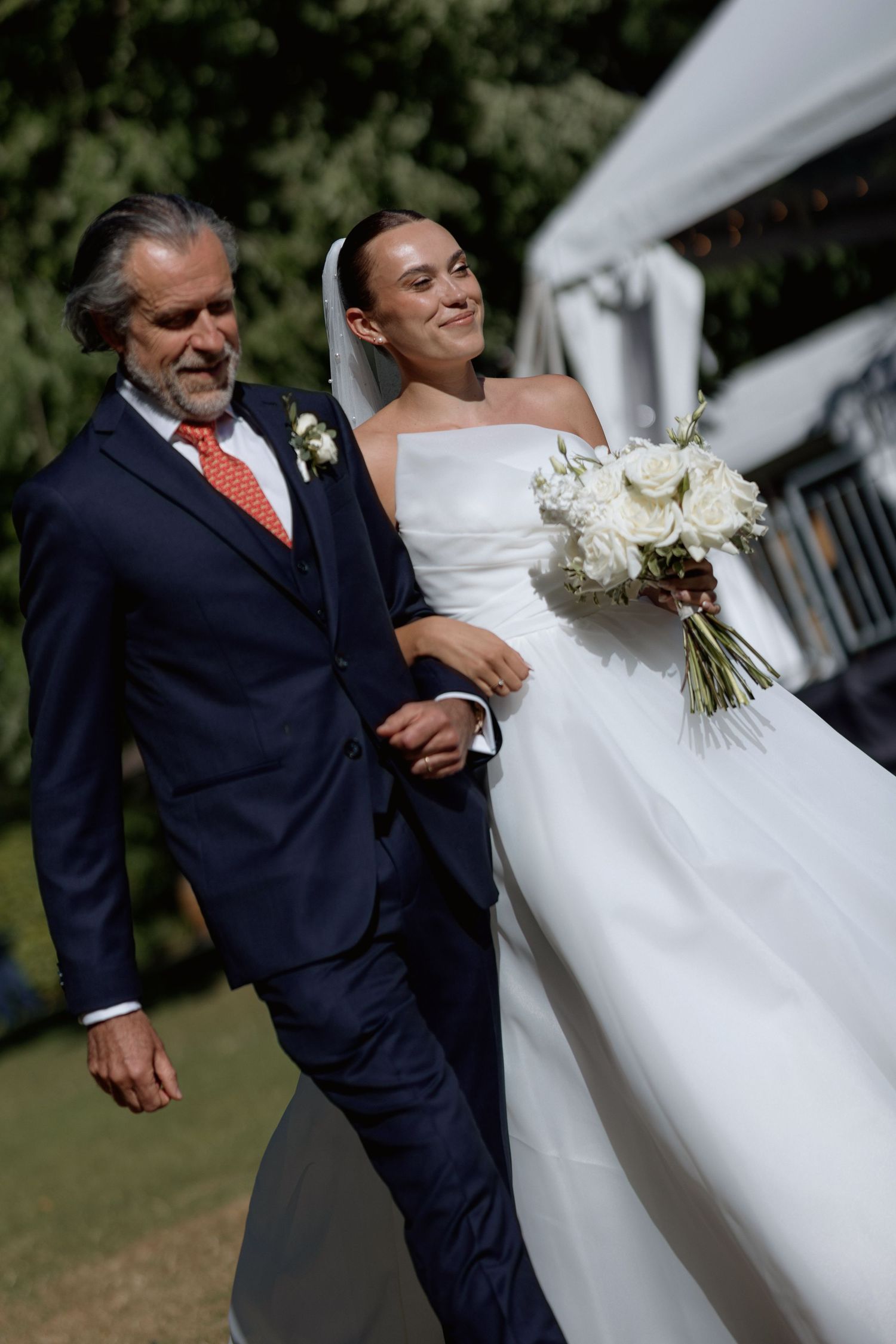 A bride in white gown walks with an escort in navy suit during an outdoor ceremony.